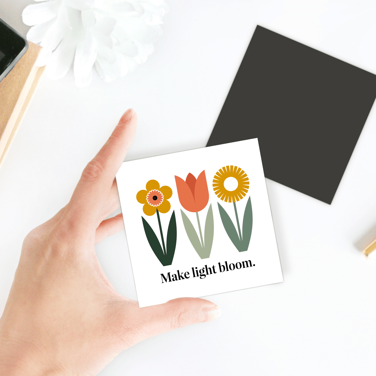 A hand holds the Make Light Bloom Magnet – Spring Floral Inspiration by Mirabilia Boutique, featuring three illustrated flowers and uplifting text—an ideal desk accent for your spring decor.