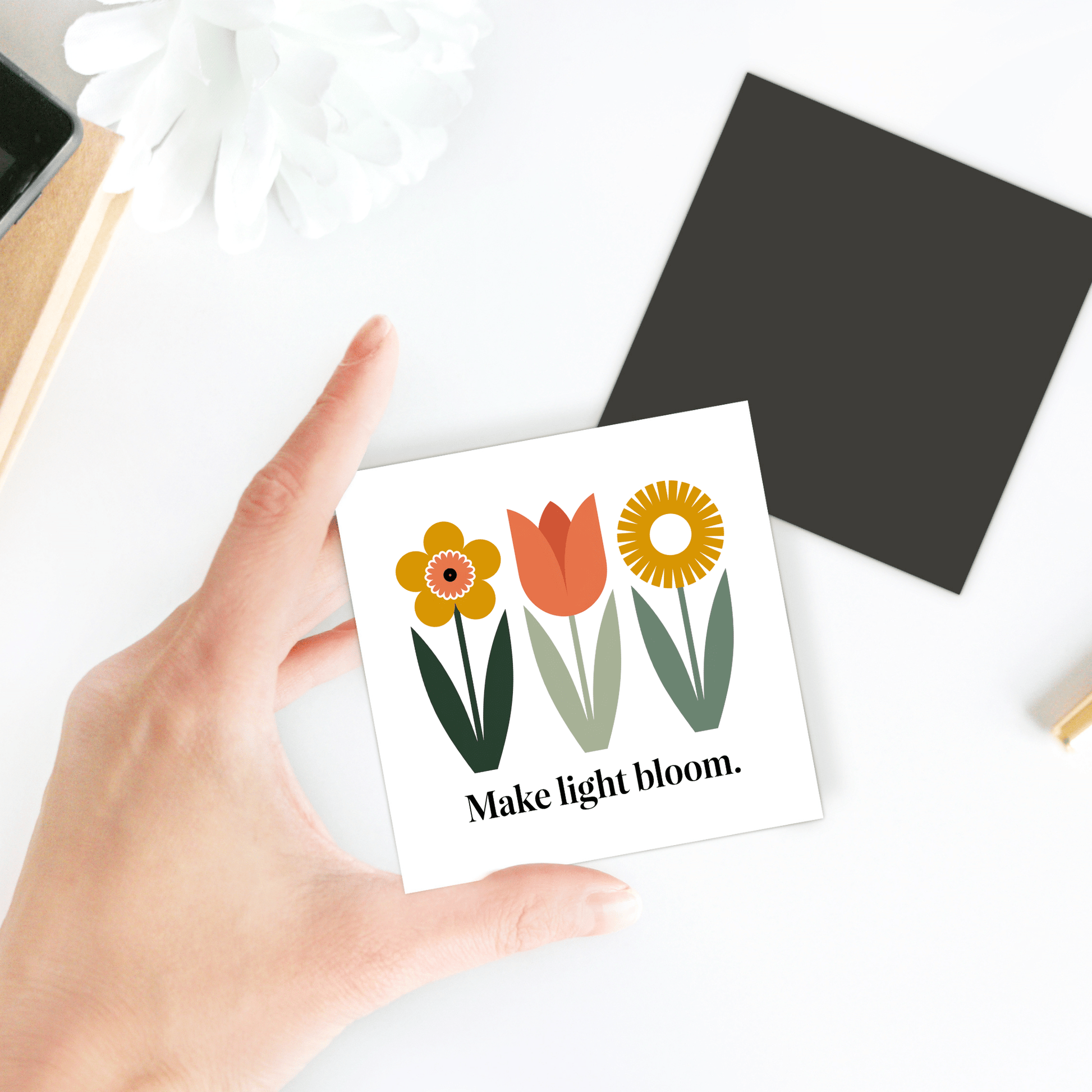 A hand holds the Make Light Bloom Magnet – Spring Floral Inspiration by Mirabilia Boutique, featuring three illustrated flowers and uplifting text—an ideal desk accent for your spring decor.