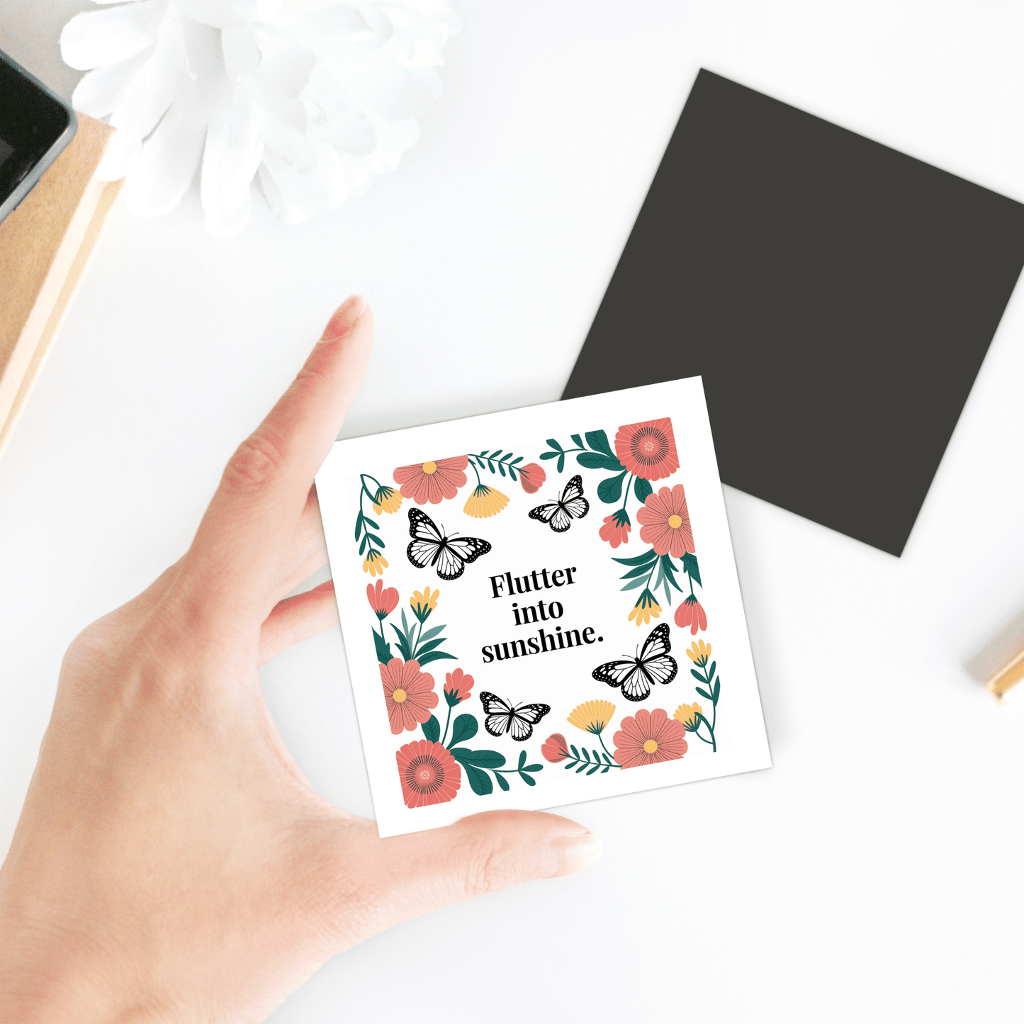 A hand holds the Flutter into Sunshine Magnet – Butterfly & Floral Joy by Mirabilia Boutique, a square card with flower and butterfly illustrations, text "Flutter into sunshine," white background, and nearby black square with a white flower.