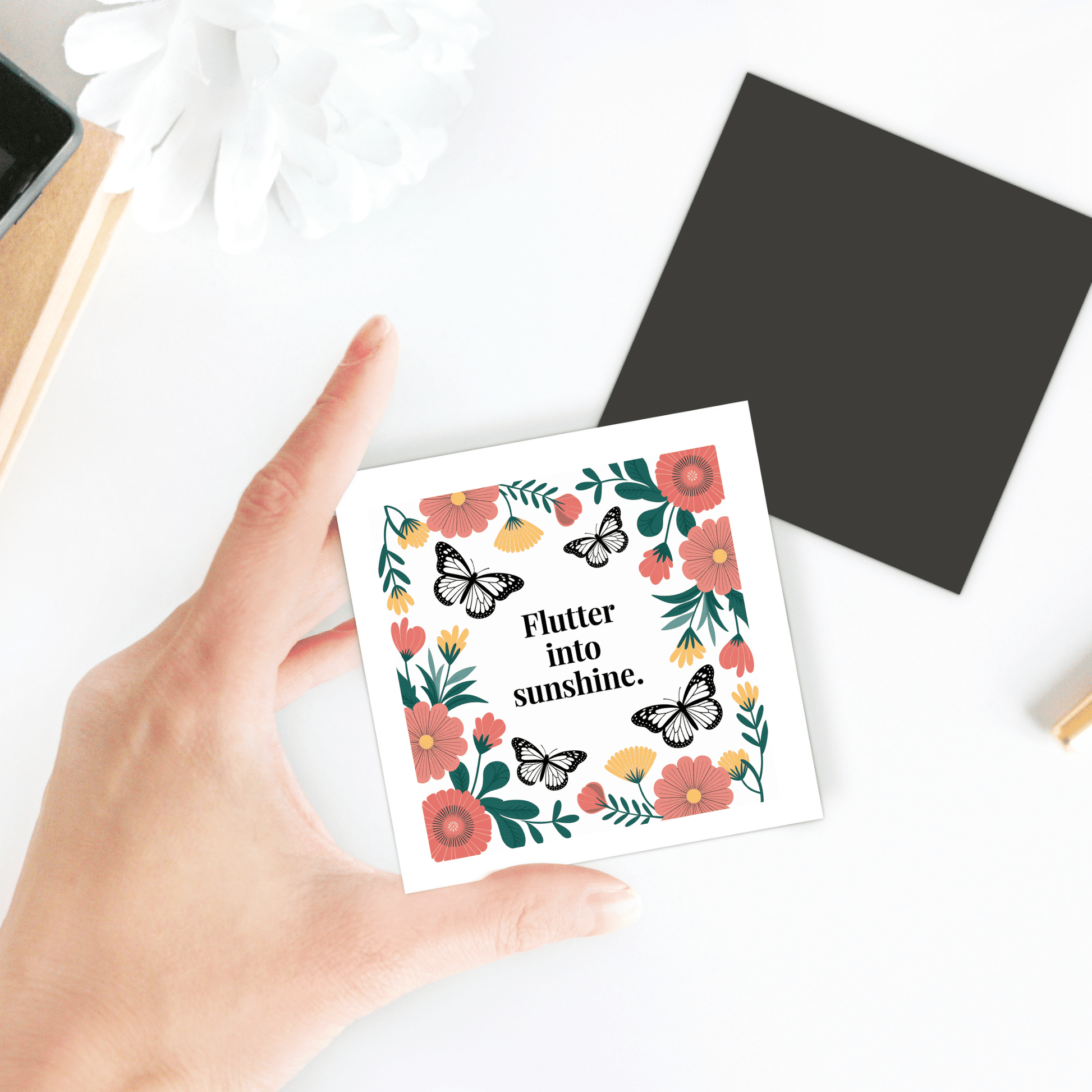 A hand holds the Flutter into Sunshine Magnet – Butterfly & Floral Joy by Mirabilia Boutique, a square card with flower and butterfly illustrations, text "Flutter into sunshine," white background, and nearby black square with a white flower.