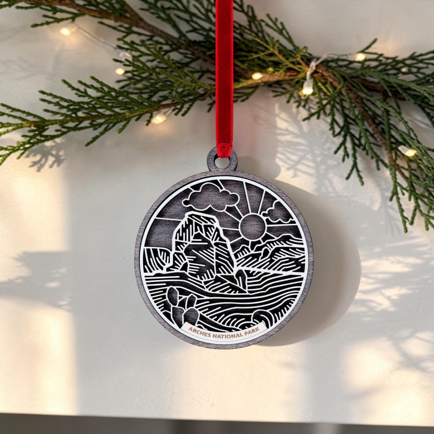 The Arches National Park Line Art Ornament by Mirabilia Boutique features a scenic landscape design in delicate line art and hangs from a vibrant red ribbon. Adorned with background greenery and string lights, this handcrafted keepsake makes the perfect addition to your collection.