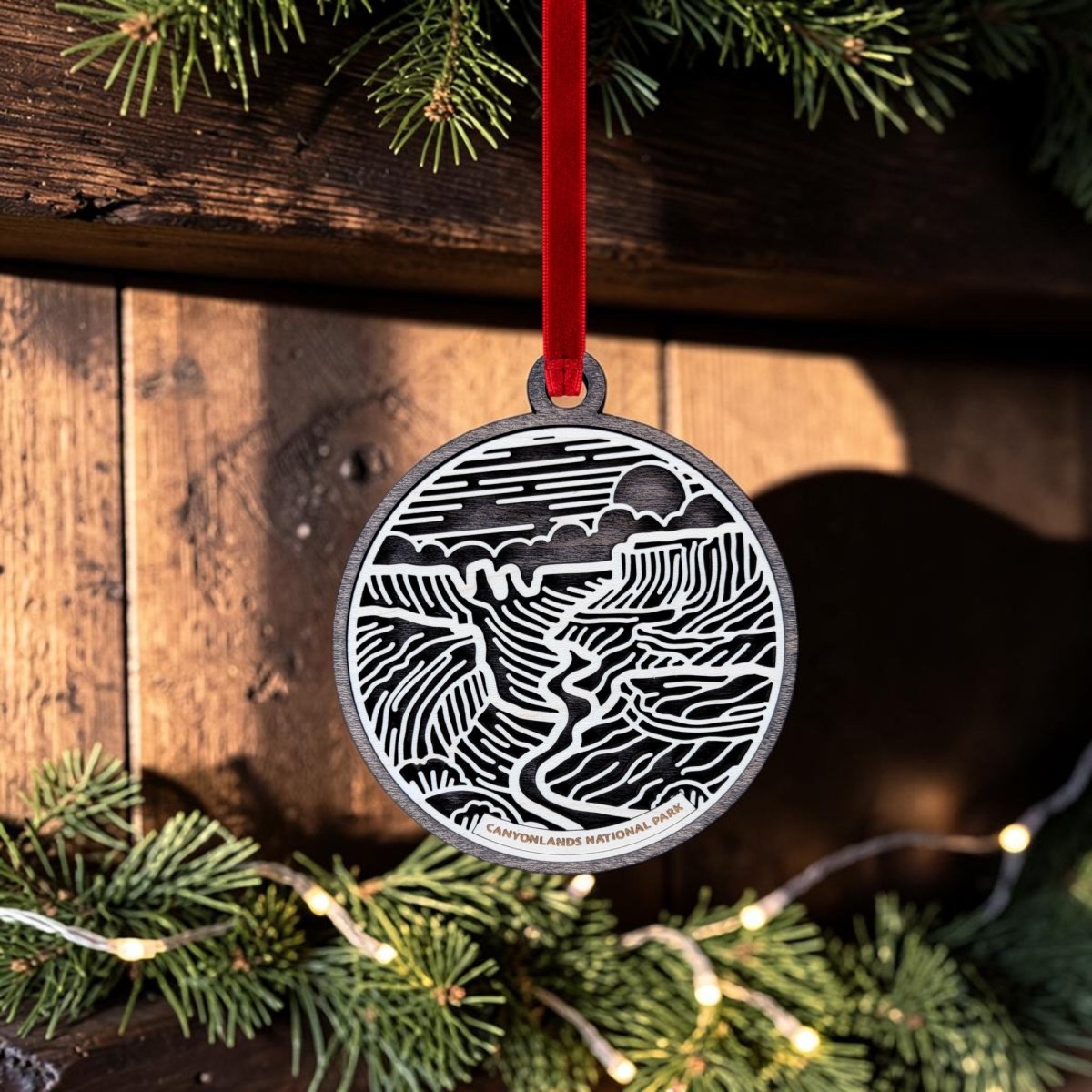 The Canyonlands National Park Christmas Ornament by Mirabilia Boutique is a round, handcrafted keepsake featuring a striking black and white landscape design. Suspended from a festive red ribbon and showcased against a wooden background adorned with pine branches and string lights, this wooden line art ornament beautifully reflects the spirit of Canyonlands National Park.