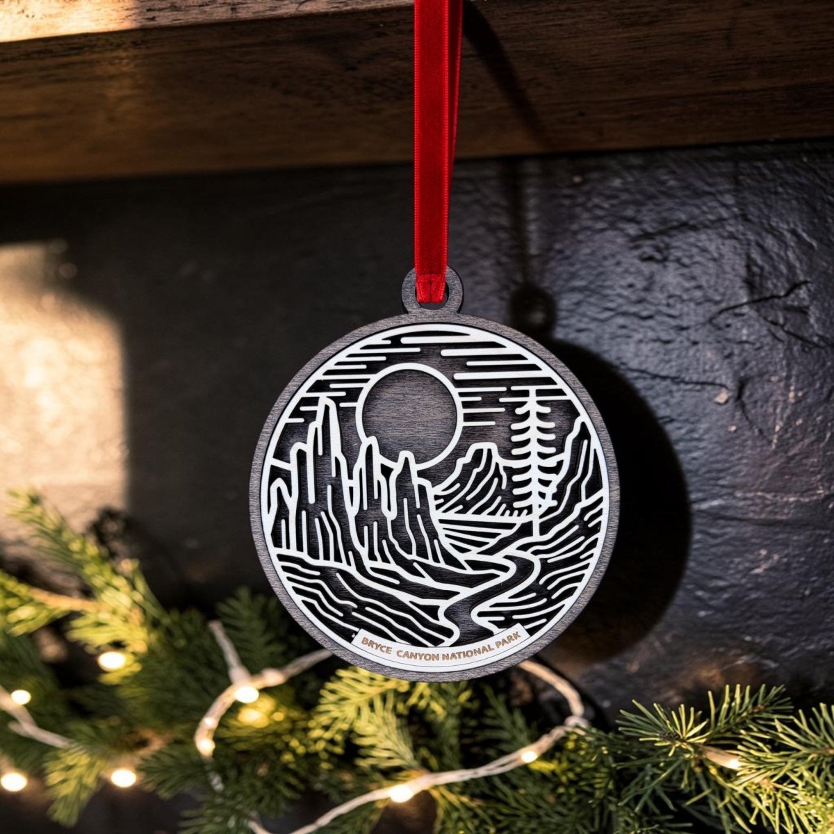 A Bryce Canyon National Park Christmas Ornament by Mirabilia Boutique, featuring a cut-out landscape design of mountains, trees, and a river in metal, hangs from a red ribbon. Labeled “Bryce Canyon National Park,” the ornament is nestled among pine branches decorated with string lights.