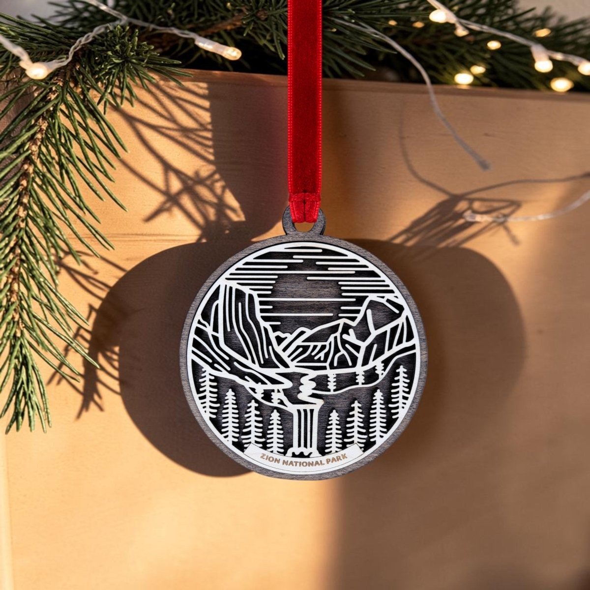 The Zion National Park Line Art Ornament by Mirabilia Boutique, featuring a delicate silver line art nature scene and "Zion National Park" text, hangs from a red ribbon on a branch adorned with pine needles and twinkling string lights in the background.
