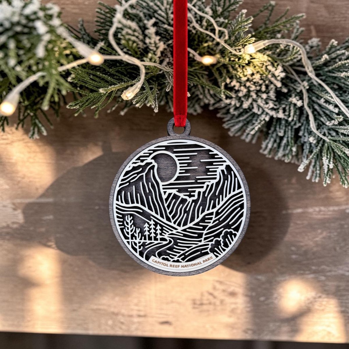 A Capitol Reef National Park Line Art Ornament by Mirabilia Boutique, featuring a wooden landscape design and the words “Capitol Reef National Park,” hangs from a red ribbon on a frosted, lit pine branch.