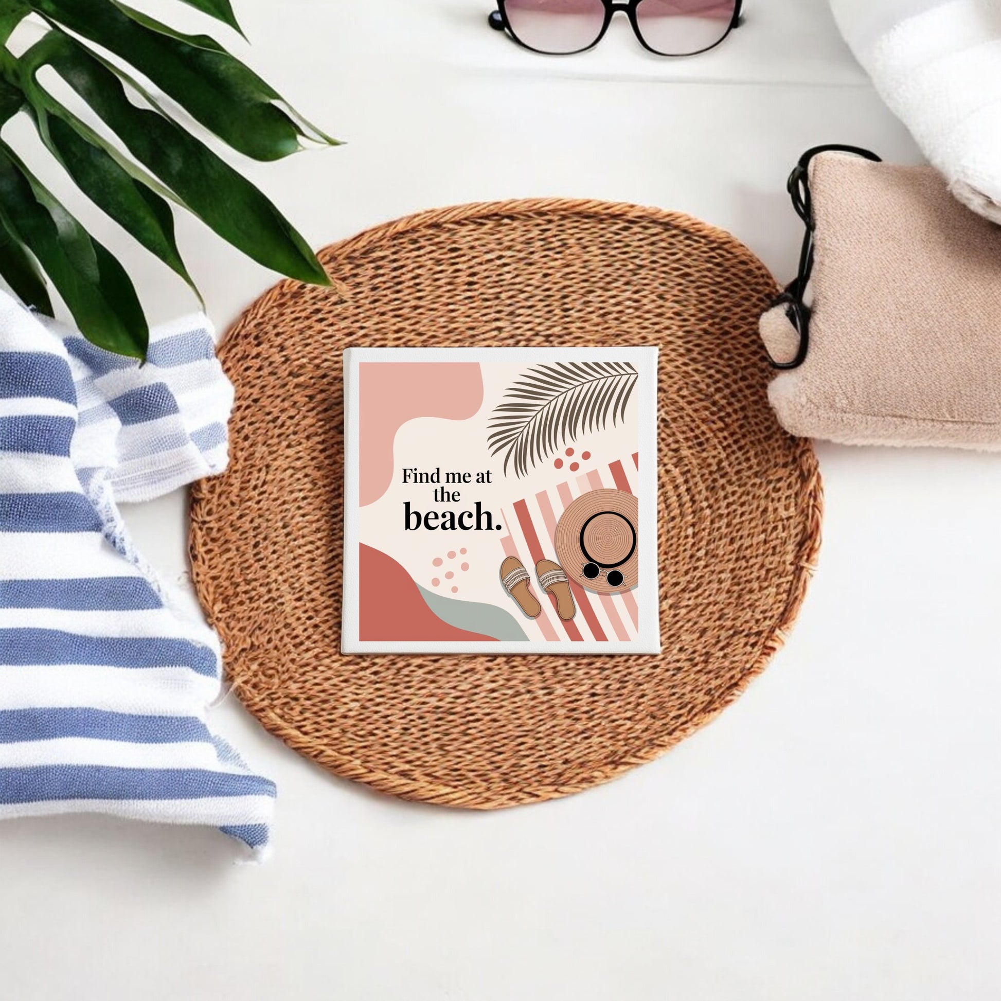 The “Find Me at the Beach Canvas – Coastal Summer Vibes” by Mirabilia Boutique rests on a wicker placemat with sunglasses, a striped towel, leaves, and a beige pouch—ideal coastal decor for your summer home.