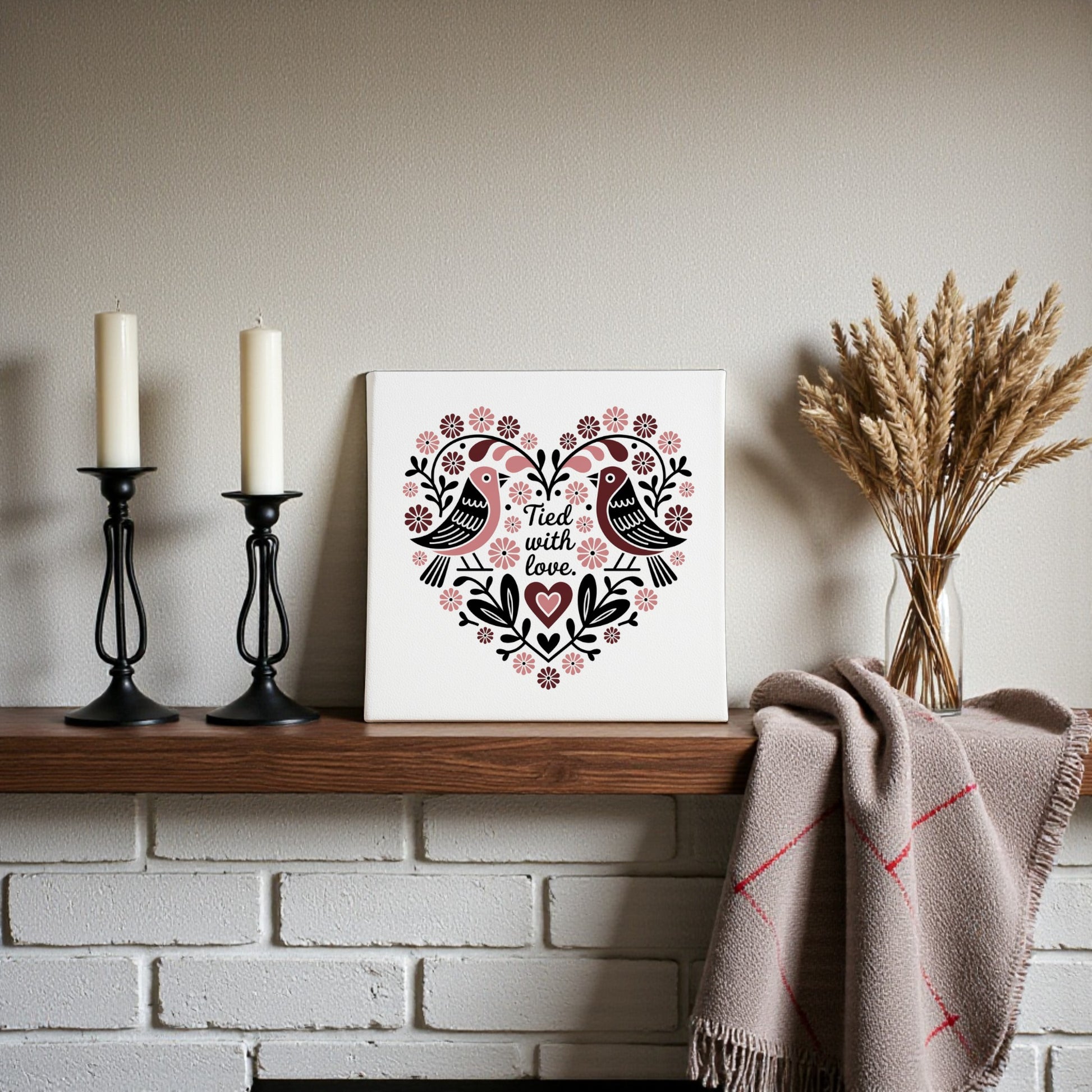 A mantel featuring two white candles in black holders, Mirabilia Boutique's "Tied With Love Folk Art Valentine Canvas," and a beige blanket with red stripes, beside a vase of dried wheat.