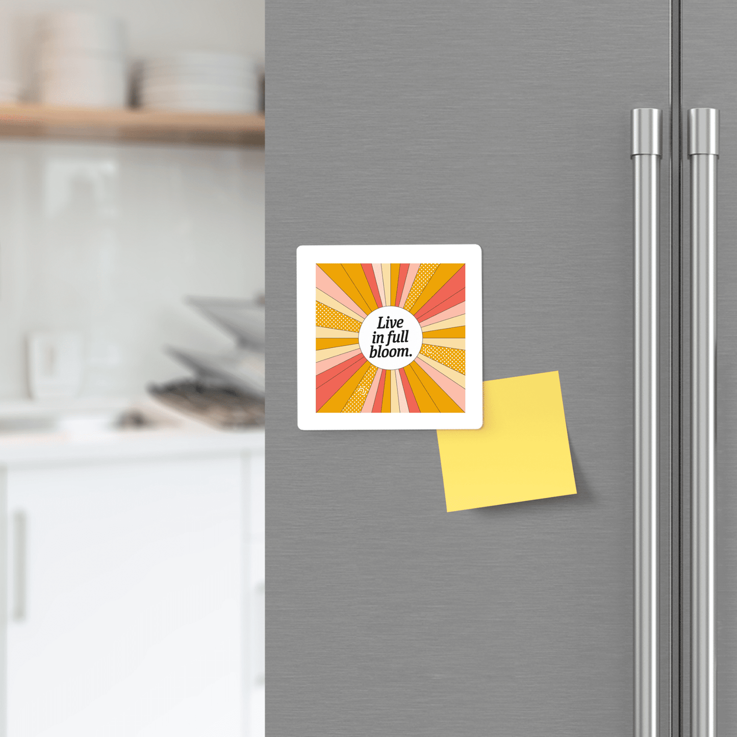 A yellow sticky note and the Mirabilia Boutique "Live in Full Bloom Magnet – Summer Sunburst Mini Art," featuring orange, yellow, and pink rays, decorate a fridge. Blurred kitchen shelves appear in the background.