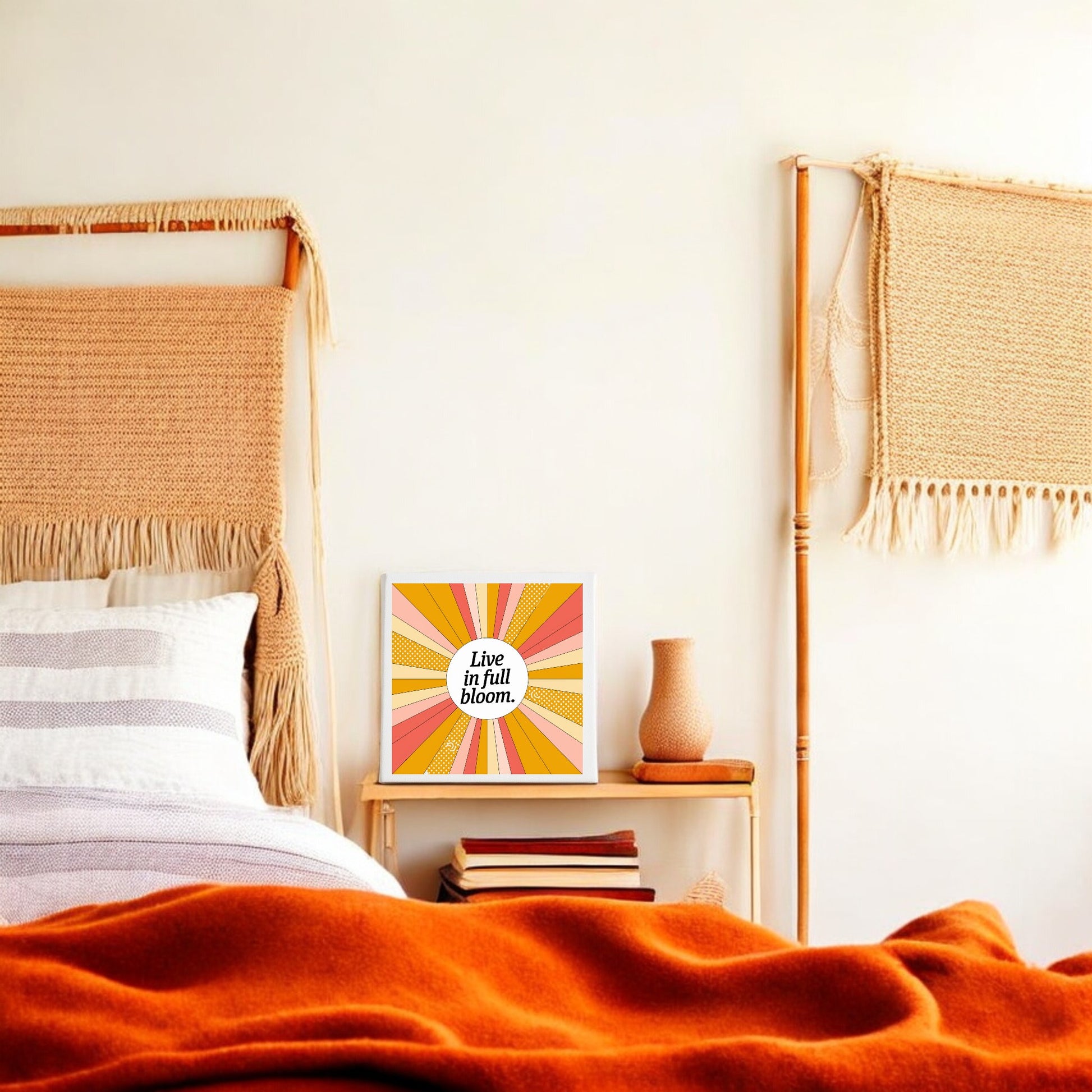 A cozy bedroom features a woven headboard, orange blanket, and nightstand with a pink vase. On the wall, Mirabilia Boutique's "Live in Full Bloom Canvas – Vibrant Summer Sunburst Art" adds an inspiring, colorful touch.