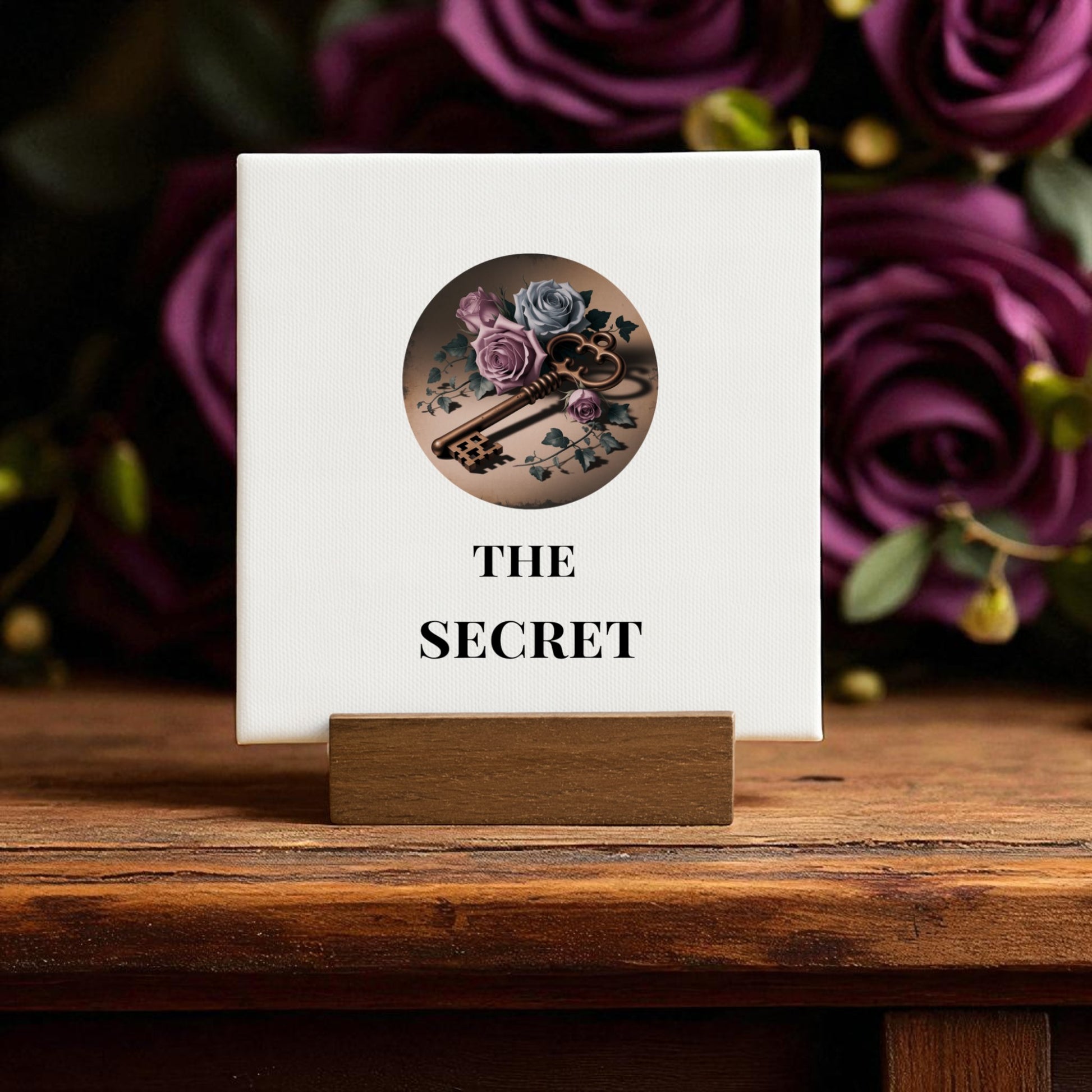 The Mini Gothic Sign by Mirabilia Boutique features a vintage skeleton key with roses and leaves encircled above "THE SECRET," set on a wooden stand with blurred purple roses—ideal Dead Regency decor for regencycore home styling.