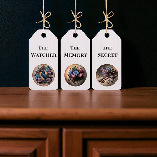 The Dead Regency Wooden Tags Set by Mirabilia Boutique includes three white wooden tags with twine, labeled "The Watcher," "The Memory," and "The Secret." Each piece features gothic floral art, perfect for dark academia decor.
