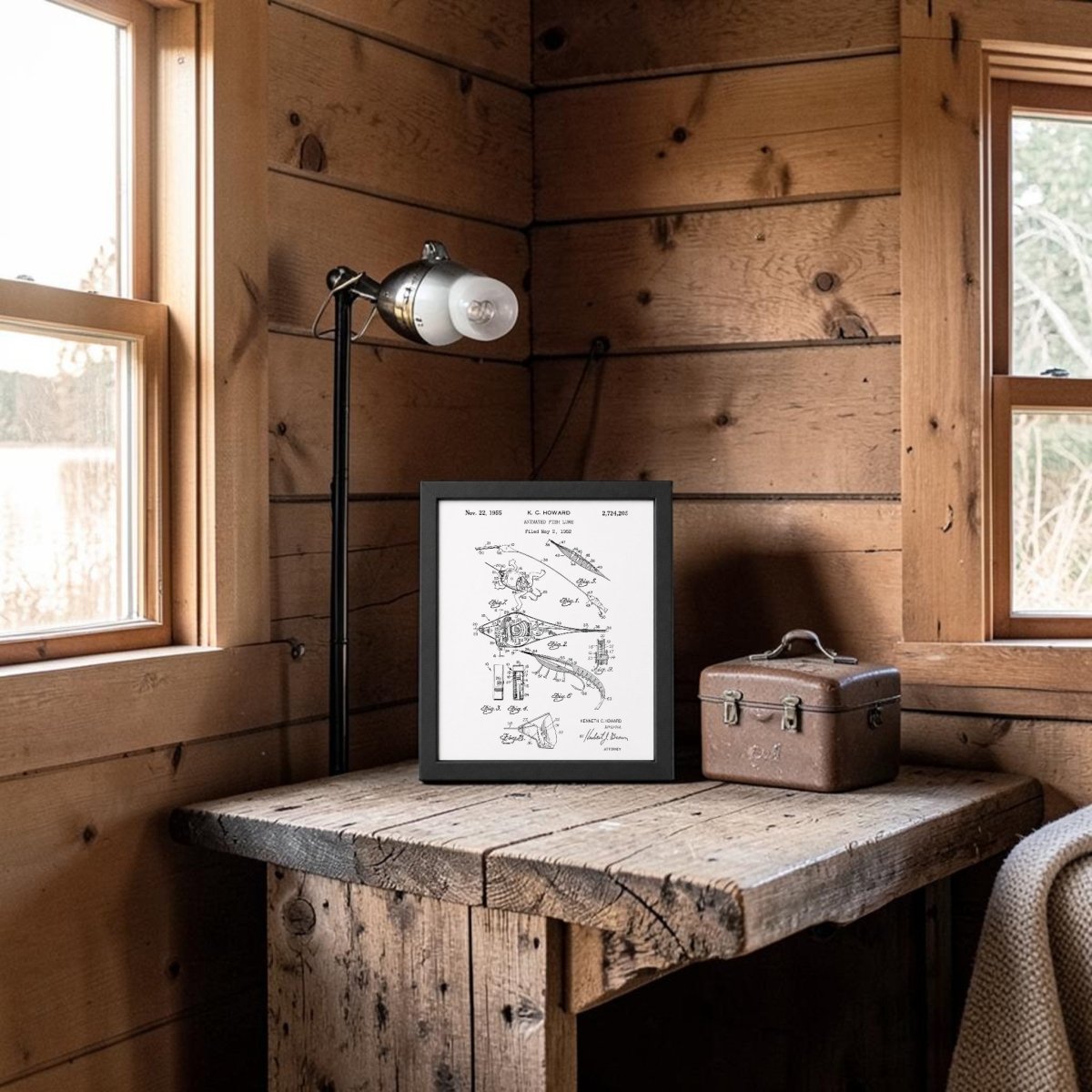 A rustic wooden corner with a window, lamp, and the Mirabilia Boutique Animated Fish Lure Patent Art Print on the table beside a small brown leather box, all nestled inside a cozy cabin.