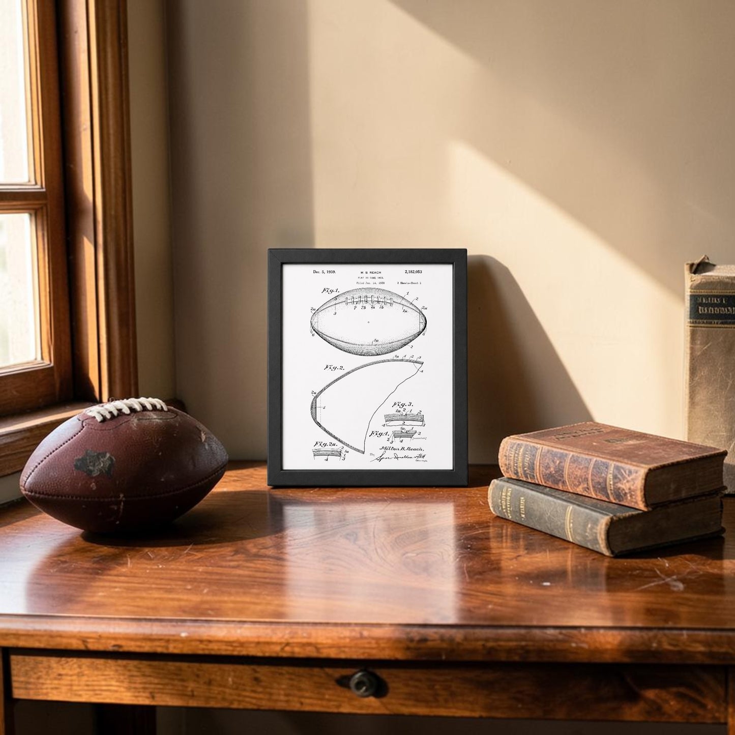 A wooden table by a window displays an old football, two vintage books, and the Mirabilia Boutique American Football Patent Art Print 8x10. Sunlight highlights the items, adding warmth and nostalgia to the scene.