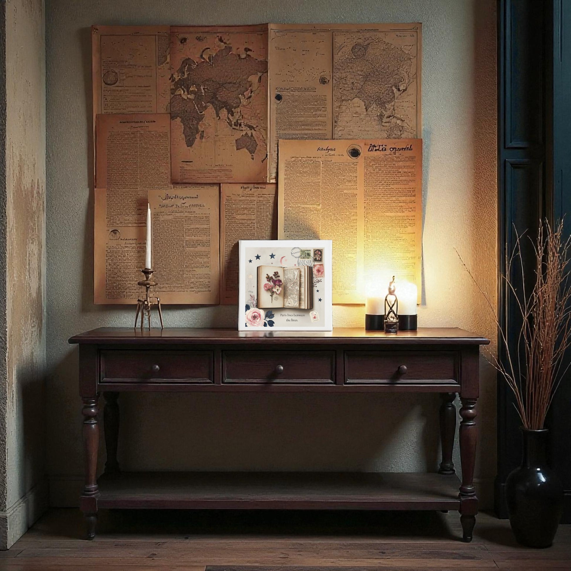 A warm scene features a vintage wooden table with three drawers against a wall decorated with old papers, maps, and the "Paris Between the Lines Canvas – Literary Floral 8x8 Art" by Mirabilia Boutique. On the table are a candle, framed collage, lantern, and dried grass in a vase.