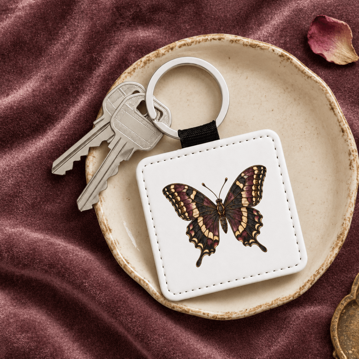 A Stained Glass Swallowtail Keychain from Mirabilia Boutique’s Velvet Ruins collection, featuring a graceful butterfly motif, rests on a ceramic dish alongside two silver keys, all arranged on luxurious burgundy velvet.