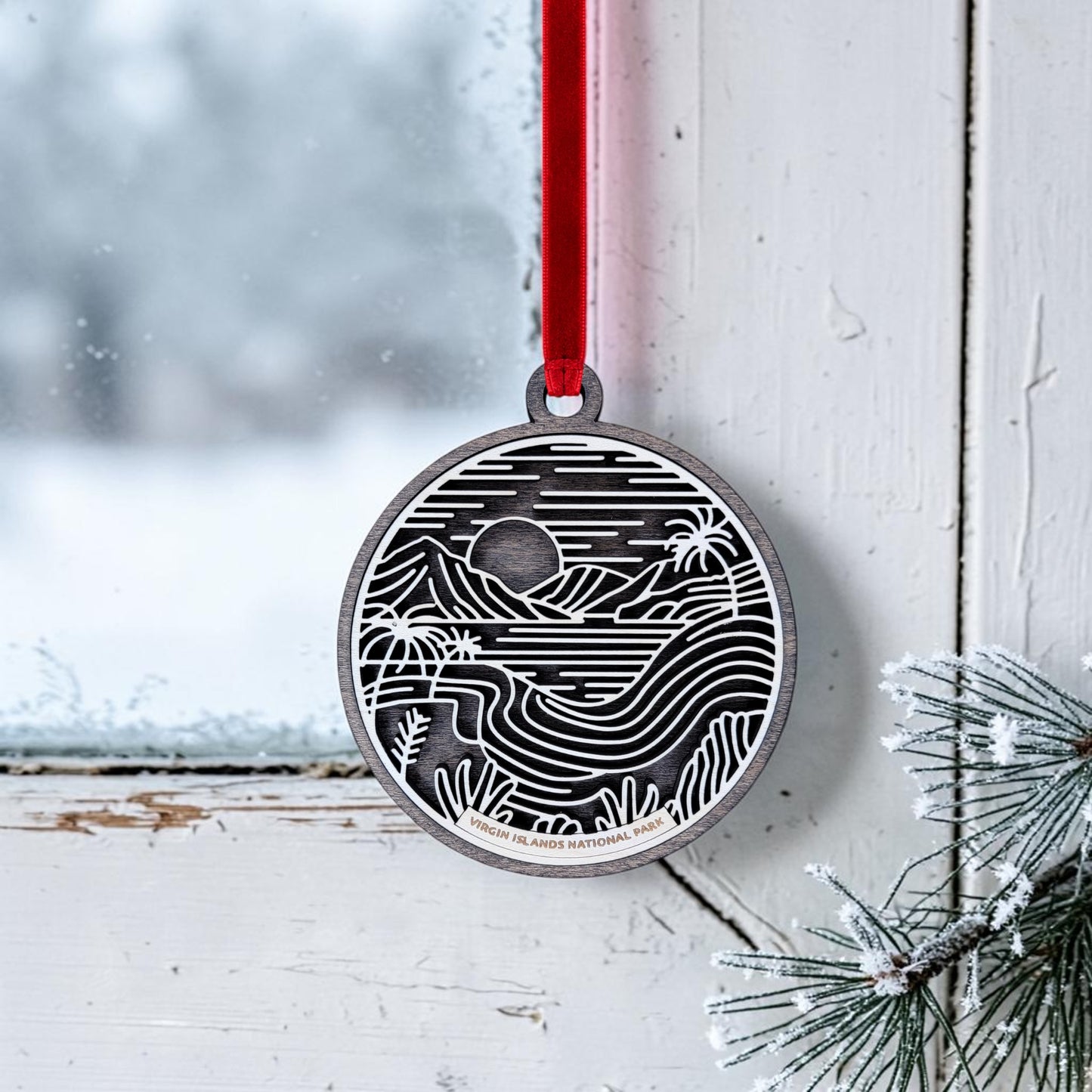 The Mirabilia Boutique Virgin Islands National Park Line Art Christmas Ornament, a round wooden piece featuring a black and white landscape design, hangs by a red ribbon on a frosted window, with snowy pine branches nearby.
