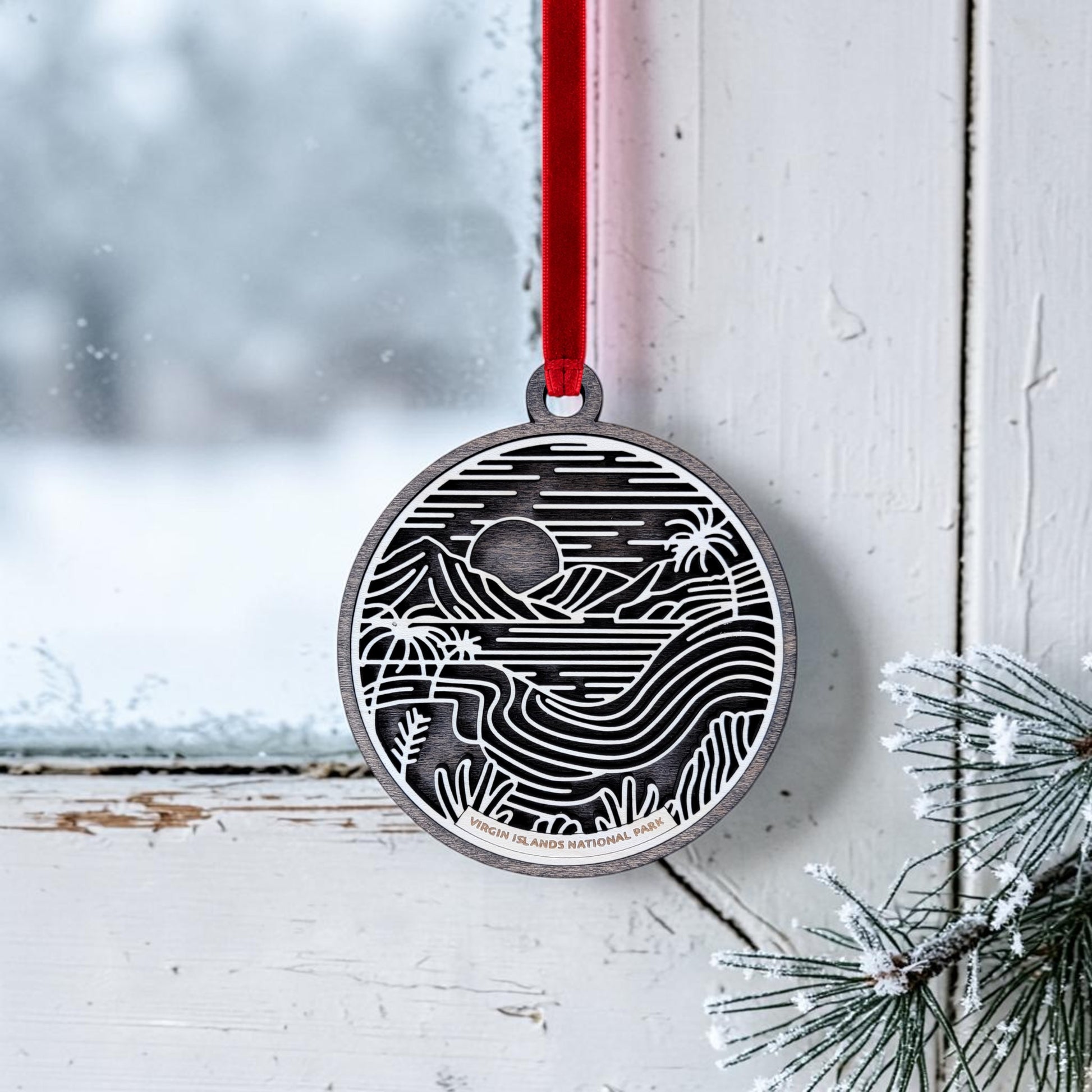 The Mirabilia Boutique Virgin Islands National Park Line Art Christmas Ornament, a round wooden piece featuring a black and white landscape design, hangs by a red ribbon on a frosted window, with snowy pine branches nearby.