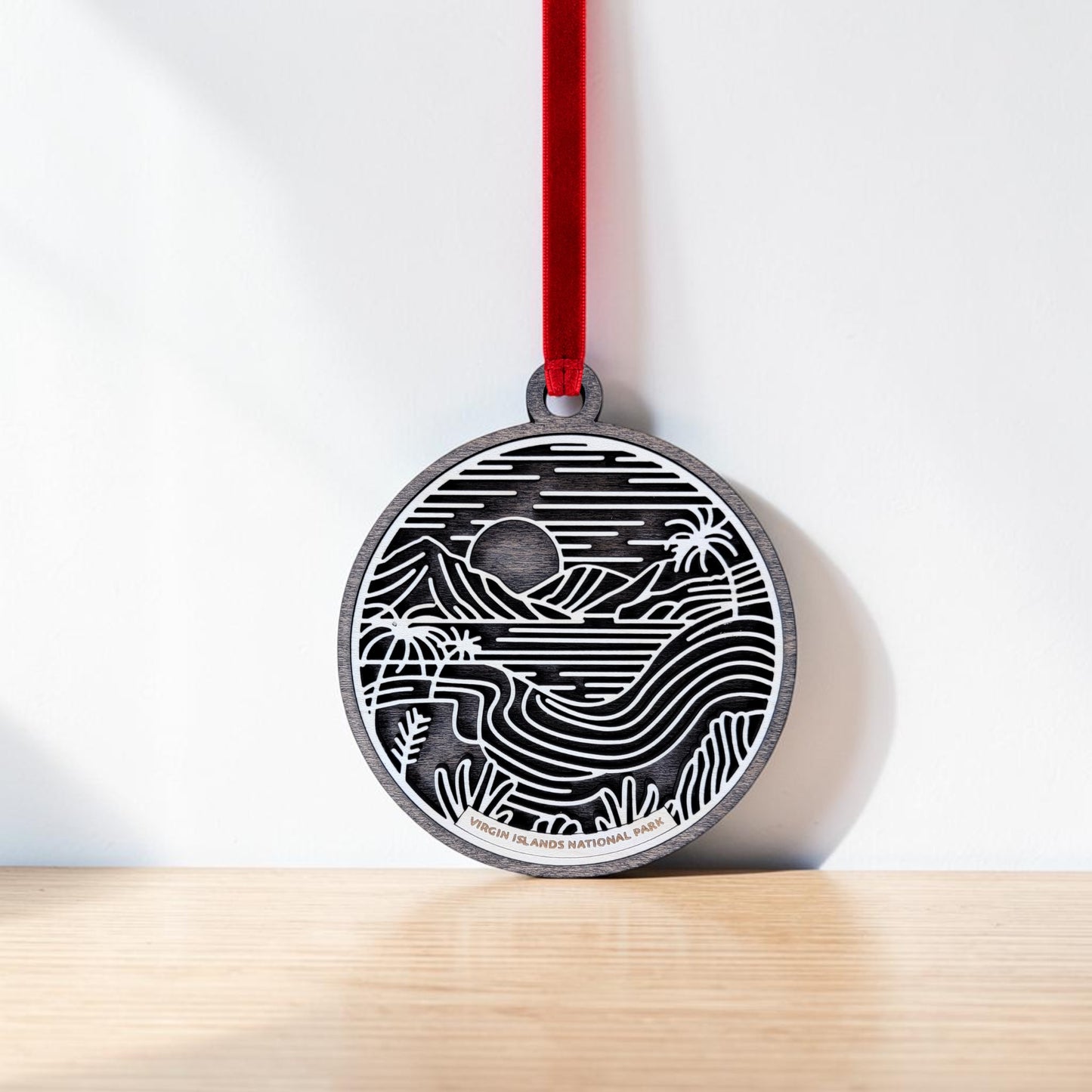 The Virgin Islands National Park Line Art Christmas Ornament by Mirabilia Boutique—a round wooden ornament featuring a black and white design of mountains, water, palm trees, and the sun—hangs on a red ribbon against a white wall, evoking Caribbean decor.