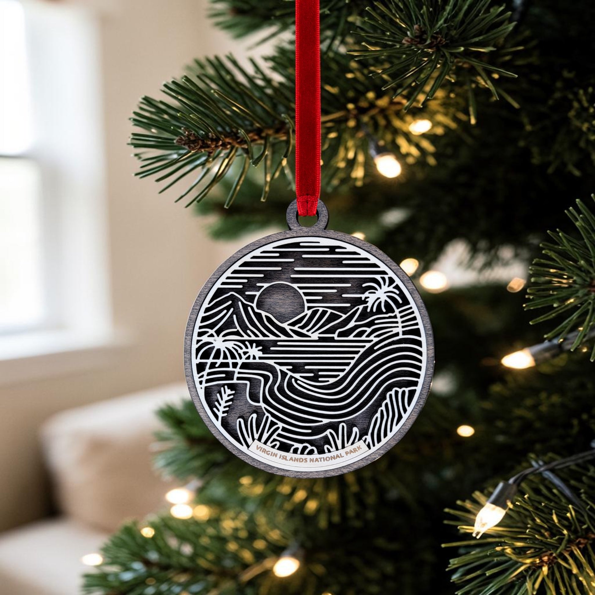 The Virgin Islands National Park Line Art Christmas Ornament by Mirabilia Boutique, featuring a black and white wooden landscape design, hangs from a red ribbon on a Christmas tree adorned with lights and set against a softly blurred background.