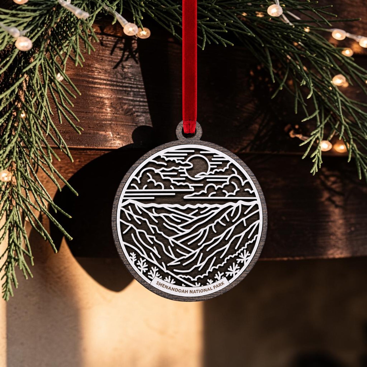 A round metal Shenandoah National Park Line Art Christmas Ornament by Mirabilia Boutique hangs from a red ribbon, showcasing a mountain and sky design. The background features evergreen branches and string lights, while the ornament displays "SHENANDOAH NATIONAL PARK" at the bottom, inspired by Blue Ridge Mountains art.