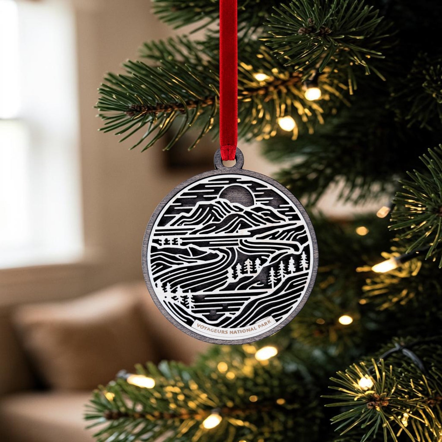 A silver oval ornament—the Voyageurs National Park Line Art Christmas Ornament by Mirabilia Boutique—hangs from a red ribbon on a lit Christmas tree, featuring a nature-inspired layered wood design and "Voyageurs National Park" amid a softly blurred indoor backdrop.
