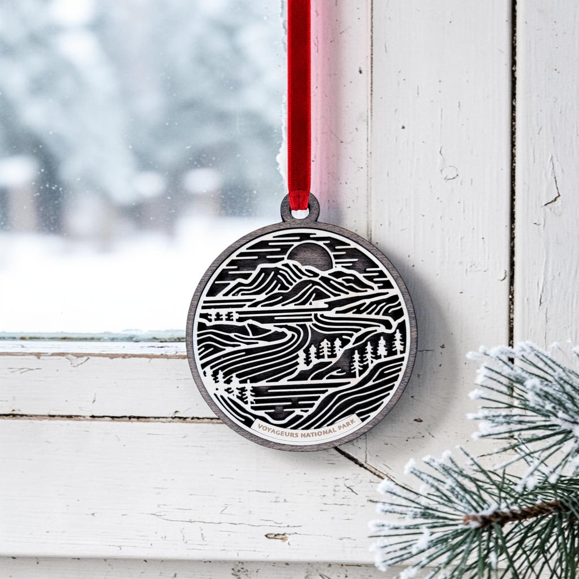 The Mirabilia Boutique Voyageurs National Park Line Art Christmas Ornament, a handcrafted wooden oval featuring a black and white landscape design, hangs from a red ribbon on a white window frame; pine branches and snow are visible outside.