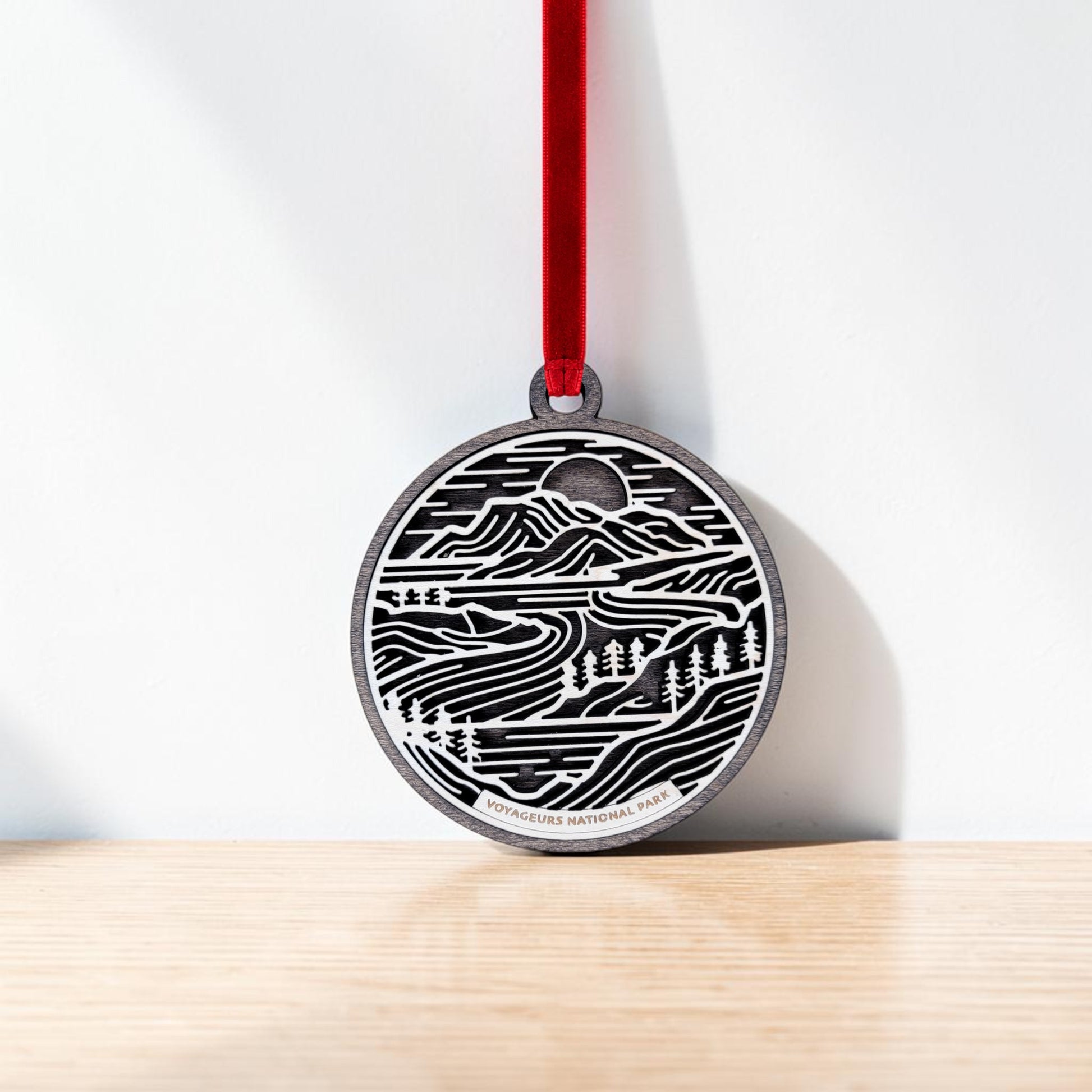 The Voyageurs National Park Line Art Christmas Ornament by Mirabilia Boutique is a handcrafted wooden decoration featuring beautifully layered wood art with mountains, trees, water, and a setting sun. Hanging by a red ribbon, the ornament displays the text "Voyageurs National Park" at the bottom and rests on a wooden surface.