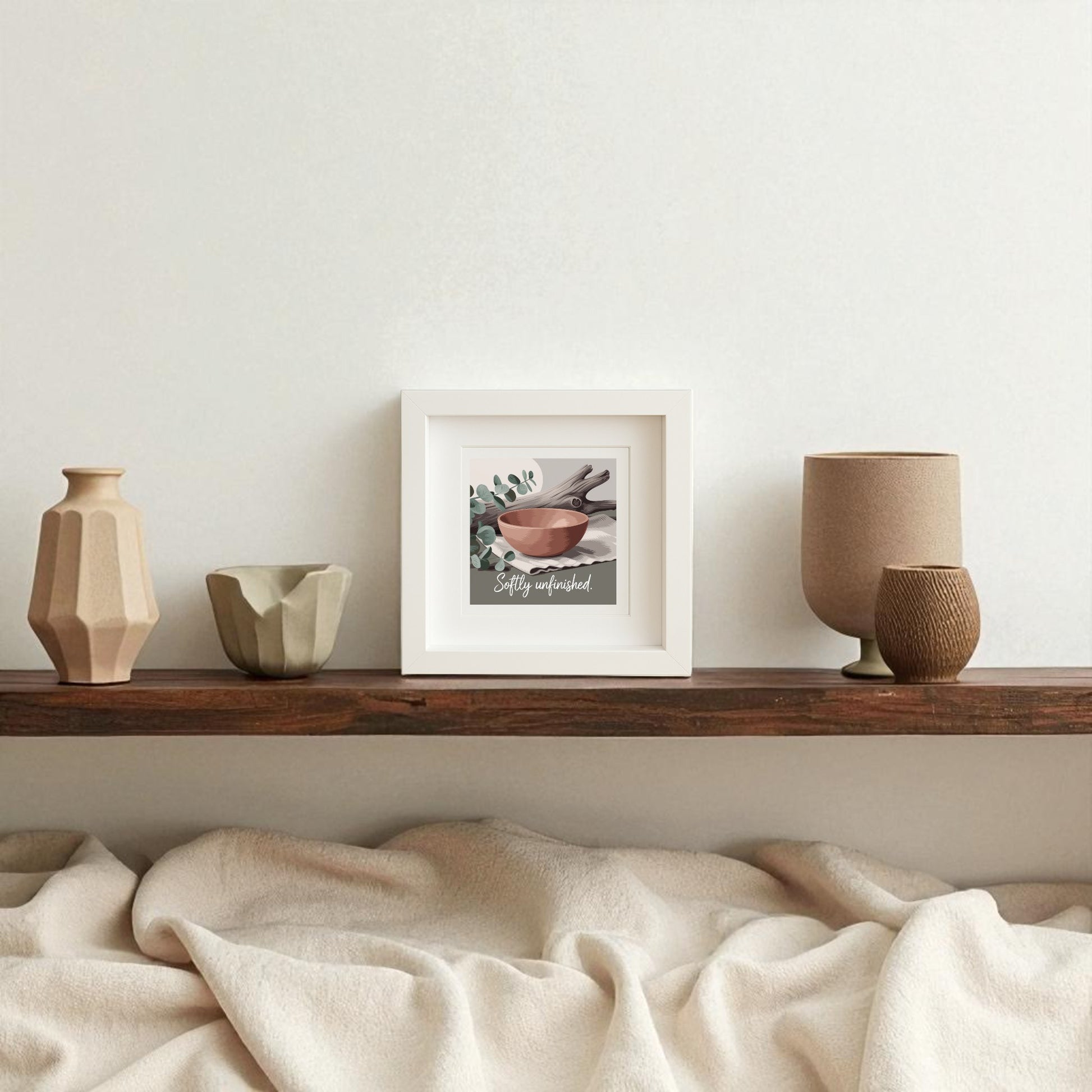 A wooden shelf holds three neutral vases and cups next to the "Softly Unfinished Shadowbox | Wabi Sabi Boho Wall Art" by Mirabilia Boutique, featuring a bowl, spoon, leaves, and "Stay wonderful." A beige blanket is draped beneath the shelf.