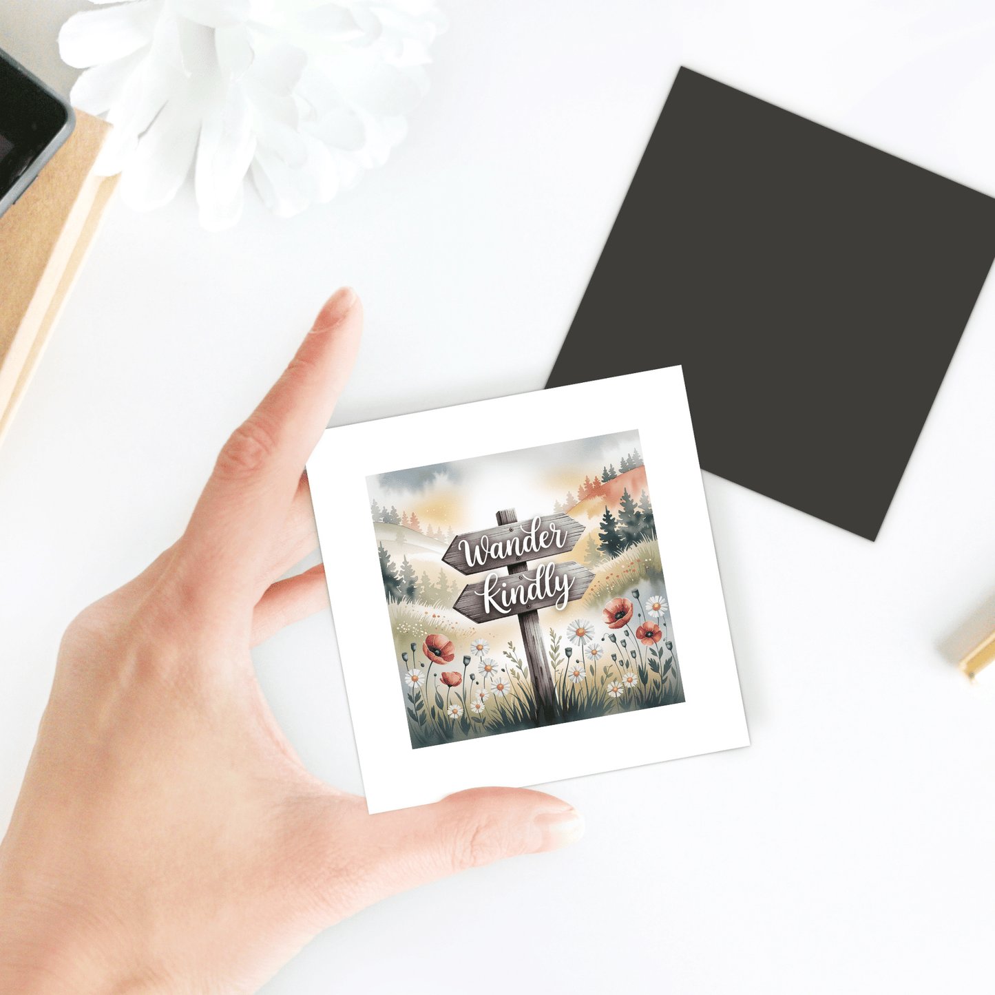 A hand holds the Wander Kindly Magnet by Mirabilia Boutique, featuring an illustrated nature scene and inspirational quote. The square magnet sits on a white desk with floral fridge magnet accents, a flower, and stationery nearby.