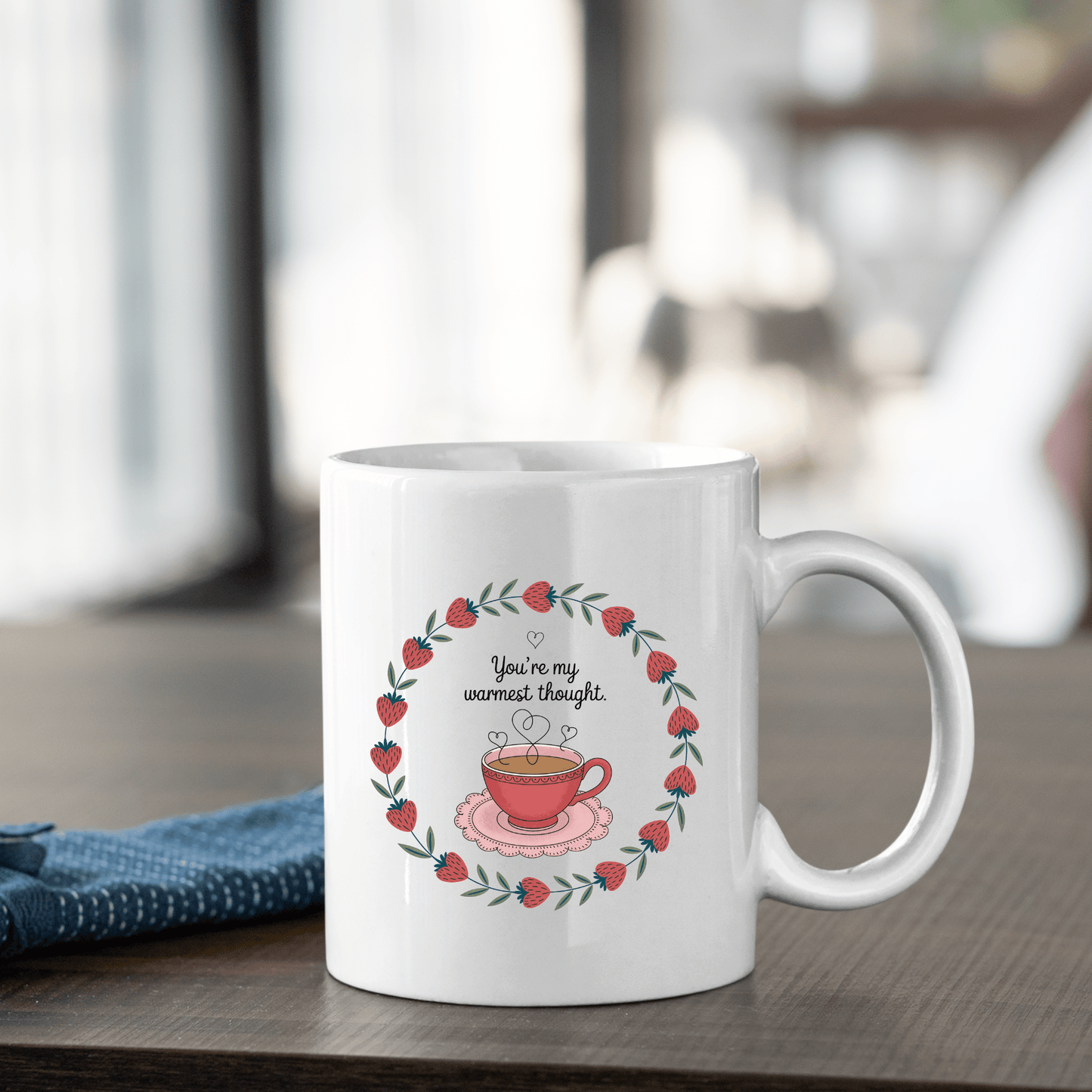 The Warmest Thought Mug – Sweet Teacup Gift Mug by Mirabilia Boutique features a steamy teacup surrounded by a strawberry wreath and the phrase “You’re my warmest thought,” perfect for cozy moments. A blue cloth rests beside this charming mug.