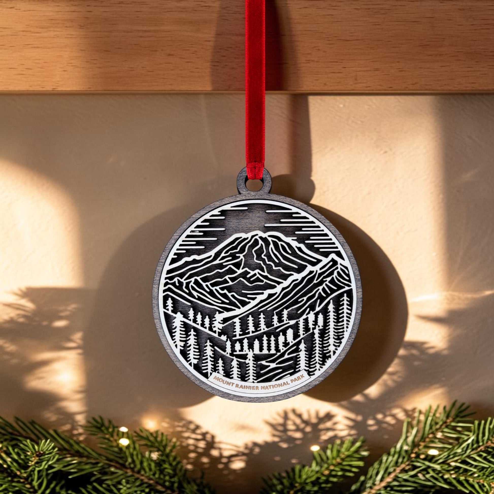 The Mount Rainier National Park Line Art Christmas Ornament by Mirabilia Boutique features an oval wooden design with a black-and-white mountain and forest illustration. It hangs from a red ribbon, with sunlight casting shadows on the wall and evergreen branches at the bottom of the image, evoking the natural beauty of Mount Rainier National Park.