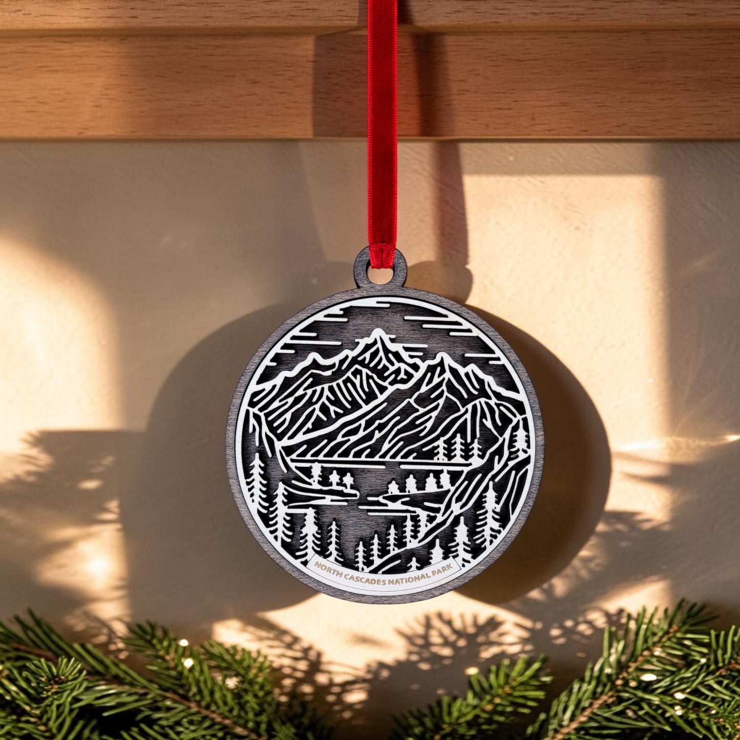 Crafted by Mirabilia Boutique, the North Cascades National Park Line Art Christmas Ornament showcases a detailed mountain and tree design with “North Cascades National Park” inscribed on it. Suspended from a red ribbon, this handcrafted metal ornament creates delicate shadows on nearby walls as sunlight streams through pine branches.