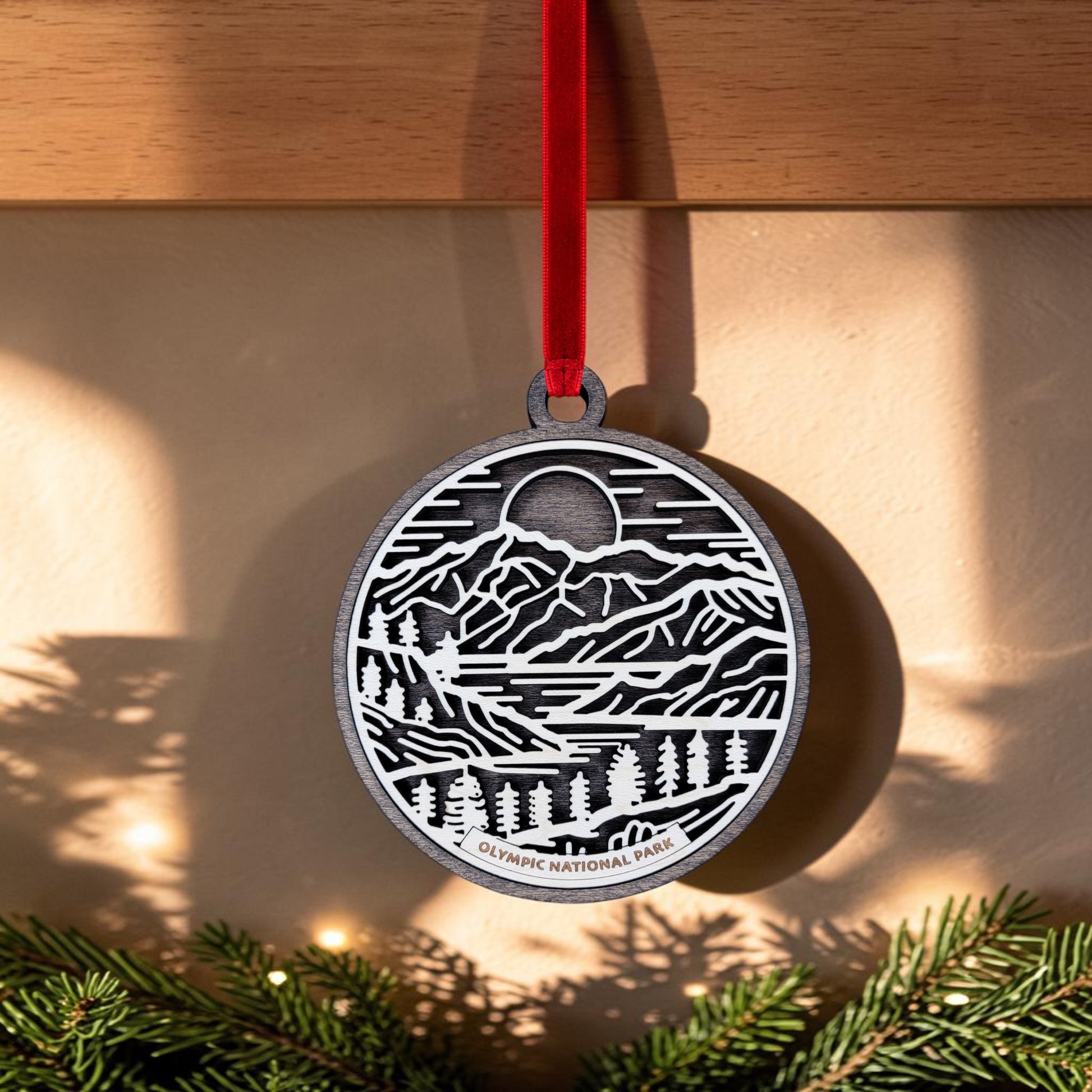 The Olympic National Park Line Art Christmas Ornament by Mirabilia Boutique is a round, handcrafted wooden keepsake adorned with a mountain, trees, and sun design. Hanging from a red ribbon, “Olympic National Park” is elegantly inscribed at the bottom. In the background, sunlight streams through evergreen branches.