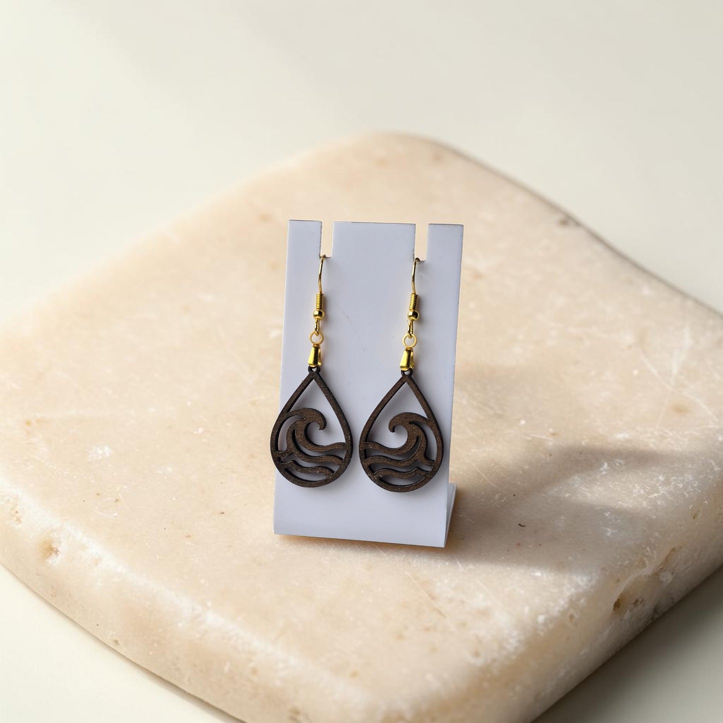 Wave Teardrop Earrings by Mirabilia Boutique, crafted from lasercut walnut wood, are displayed on a white stand atop a light stone surface. Ocean-inspired design and gold hooks add elegant charm to any outfit.
