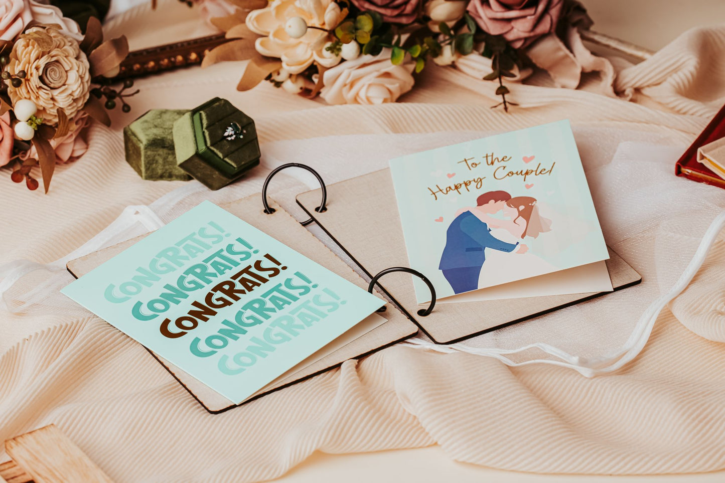 Mirabilia Boutique’s Wedding Card Keepsake Binder | Personalized Memory Book displays pastel cards—one says "CONGRATS!" and another shows an embracing couple with "To the Happy Couple!"—a lovely wedding keepsake amid flowers and fabric.