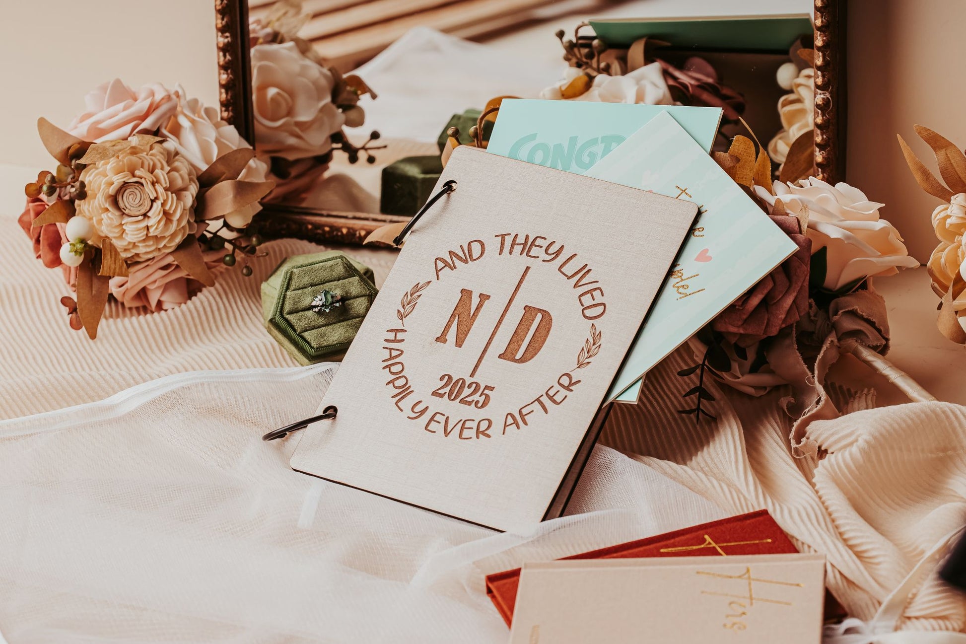 The Wedding Card Keepsake Binder | Personalized Memory Book by Mirabilia Boutique, featuring "And They Lived Happily Ever After N/D 2025" on the cover, is displayed on fabric with flowers, a green ring box, cards, and a decorative mirror.