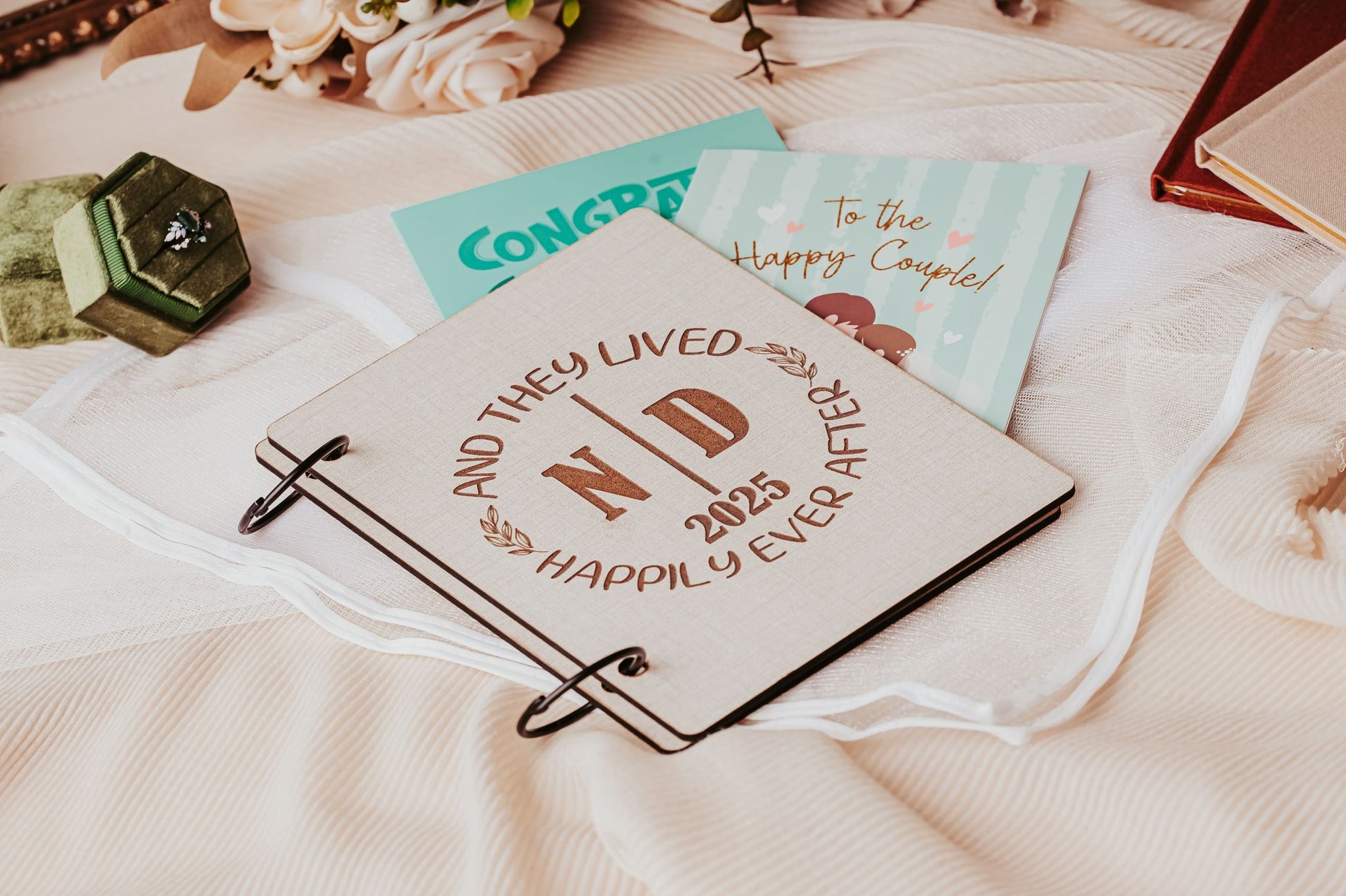 A wooden sign with “And They Lived Happily Ever After N D 2025” rests on pink fabric beside a green ring box, congratulatory cards, flowers, and the Mirabilia Boutique Wedding Card Keepsake Binder | Personalized Memory Book.