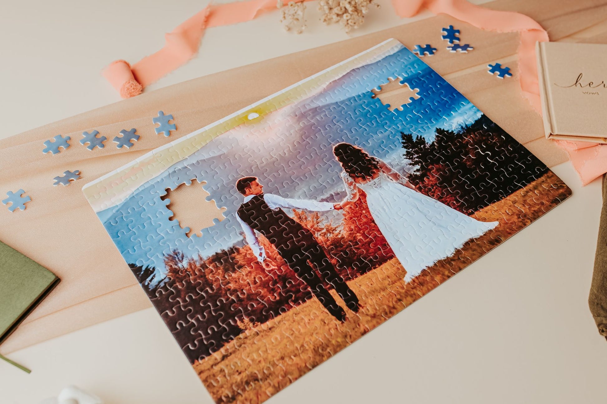 A partially finished Mirabilia Boutique Custom Wedding Jigsaw Puzzle | 300-Piece Photo Keepsake displays a couple in wedding attire holding hands at sunset, with blue-edge pieces missing and loose pieces on a peach cloth.