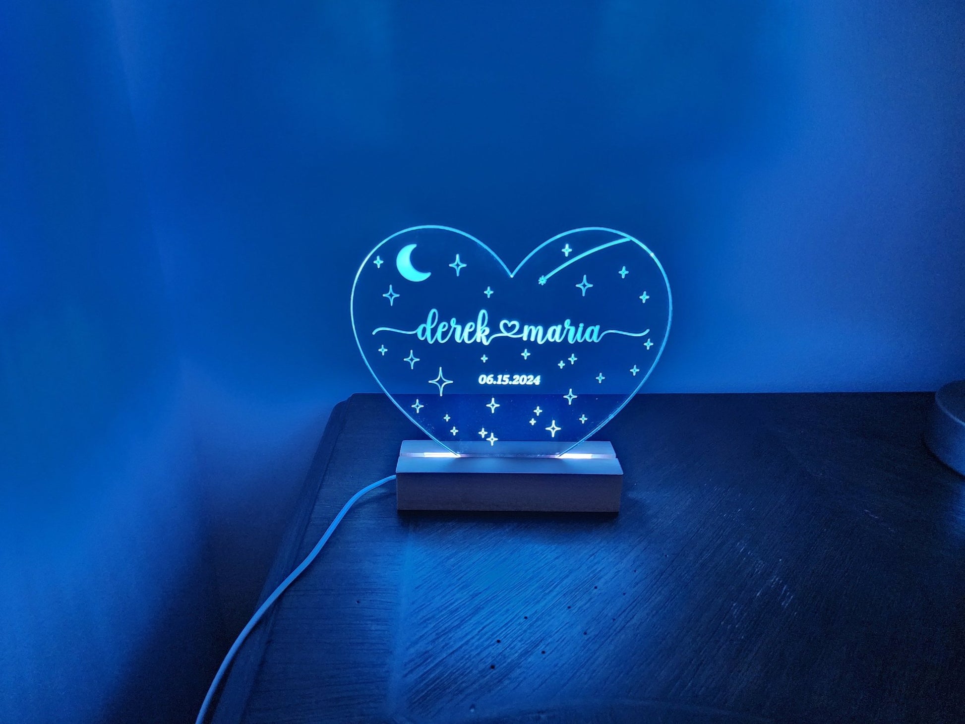 Introducing the Personalized Wedding Date Night Light Custom Keepsake by Mirabilia Boutique: A heart-shaped LED night light with "derek ♥ maria" and "06.15.2024" beautifully engraved on it. This elegant gift emits a soothing blue glow and features whimsical star and moon designs. Mounted on a wooden base, it stands out perfectly against a blue-lit wall, making it an ideal memento for any wedding celebration.