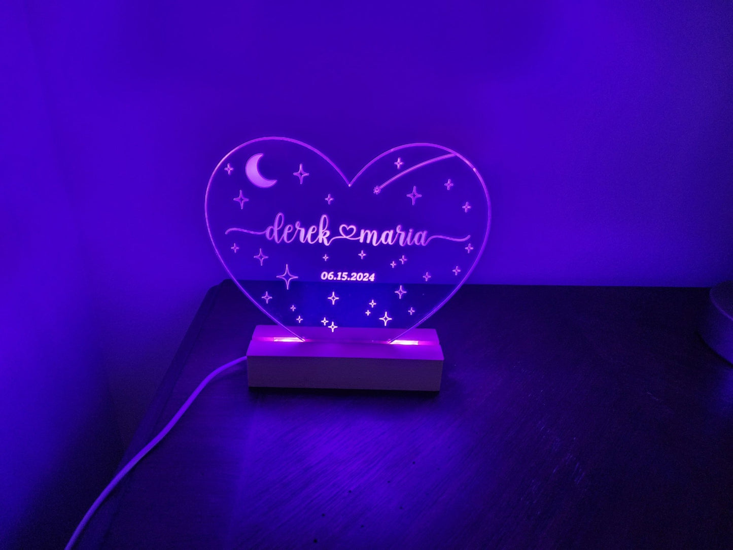 A heart-shaped LED night light from Mirabilia Boutique, called the "Personalized Wedding Date Night Light Custom Keepsake," features star and moon designs along with the names "Derek & Maria" and their wedding date "06.15.2024." This customized wedding gift emits a purple glow, resting on a dark surface with a power cord visible on the left side.