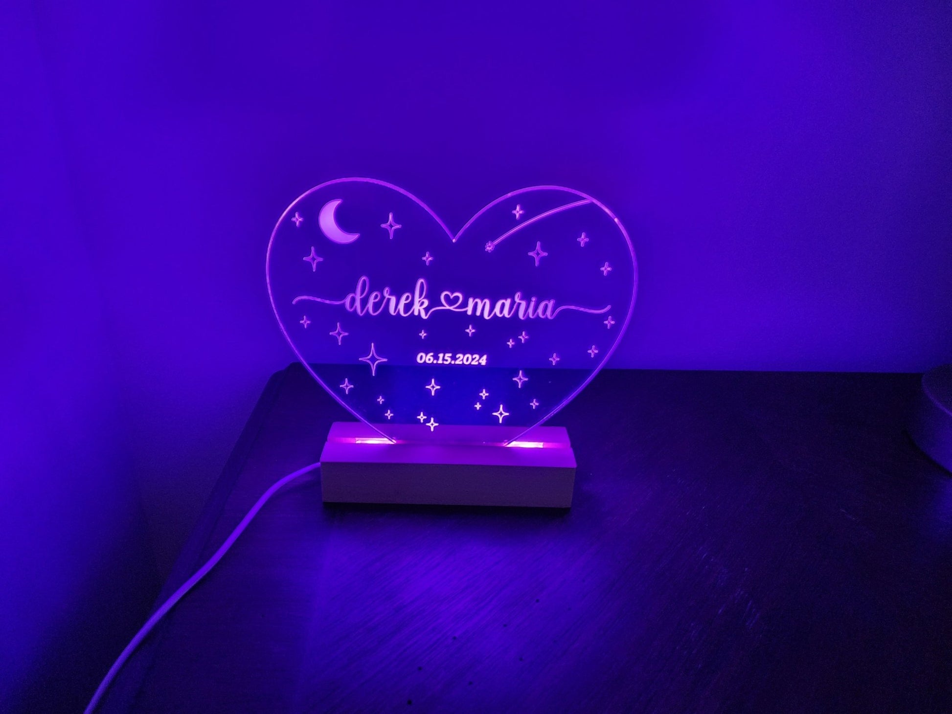 A heart-shaped LED night light from Mirabilia Boutique, called the "Personalized Wedding Date Night Light Custom Keepsake," features star and moon designs along with the names "Derek & Maria" and their wedding date "06.15.2024." This customized wedding gift emits a purple glow, resting on a dark surface with a power cord visible on the left side.