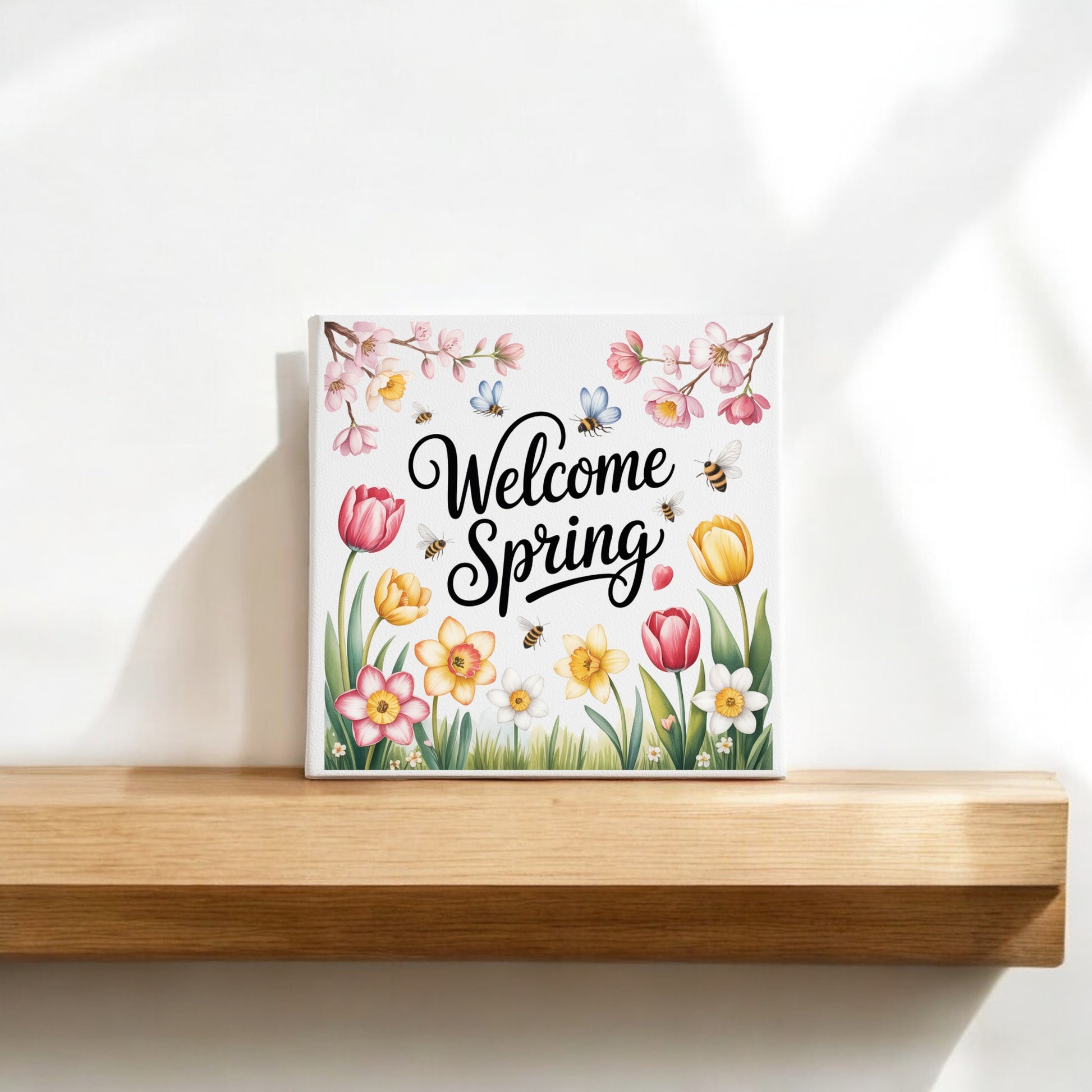 The Mirabilia Boutique Welcome Spring Canvas – 8x8 Floral Wall Art features colorful flowers, bees, and butterflies for cheerful seasonal decor, with sunlight casting gentle shadows wherever it’s displayed.