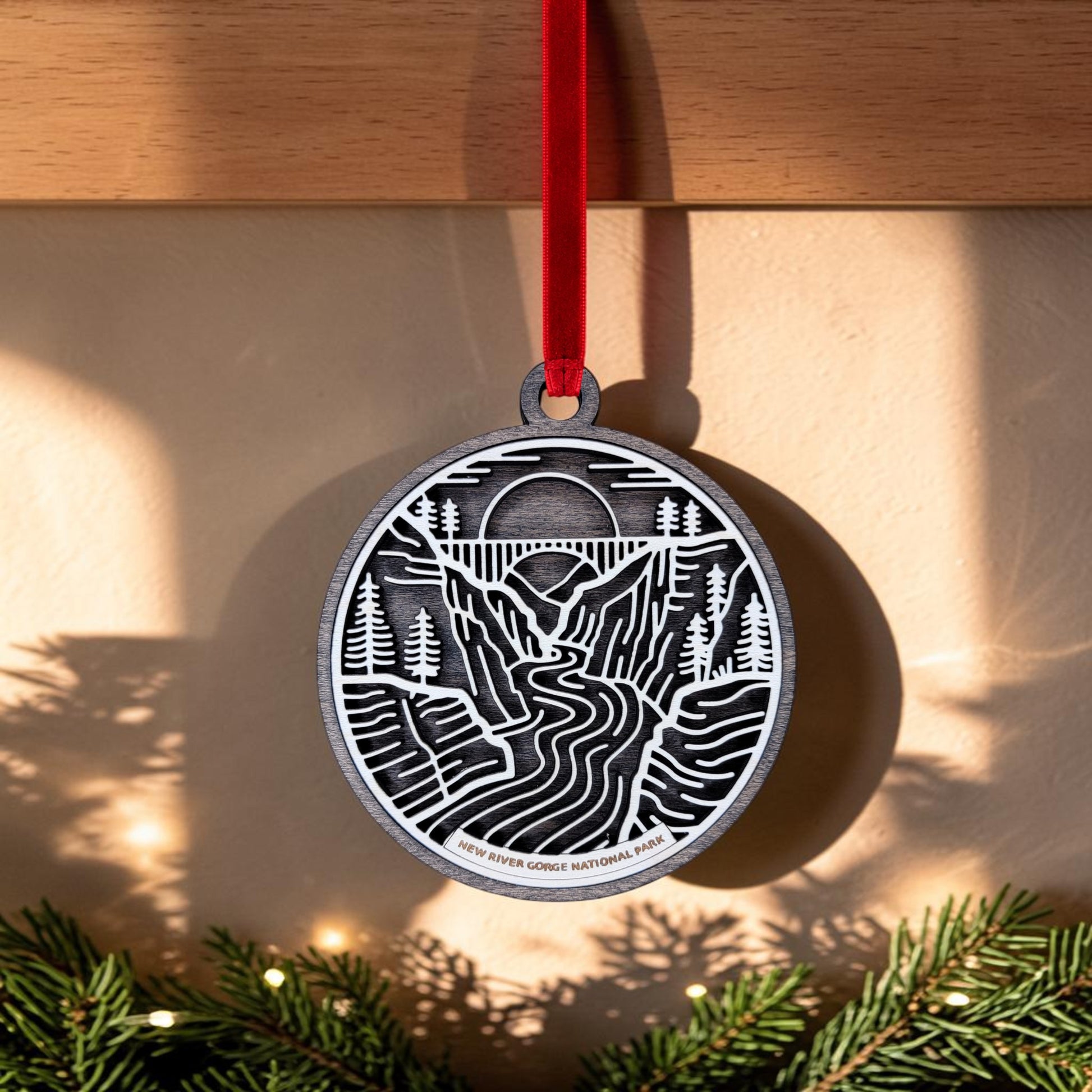 The New River Gorge National Park Line Art Christmas Ornament by Mirabilia Boutique is an oval wooden ornament featuring a cutout landscape design with mountains, trees, and the iconic New River Gorge Bridge arching over a winding river. It hangs from a red ribbon, with "New River Gorge National Park" etched at the bottom and pine branches elegantly framing the lower edge.