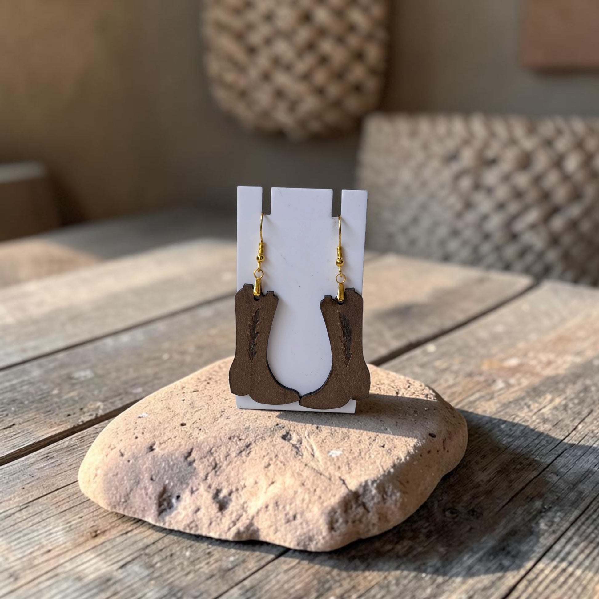 The Cowboy Boot Earrings | Lasercut Walnut Wood by Mirabilia Boutique are shown on a white stand atop a rock and rustic wooden table, with textured decor and warm lighting highlighting this unique western jewelry.