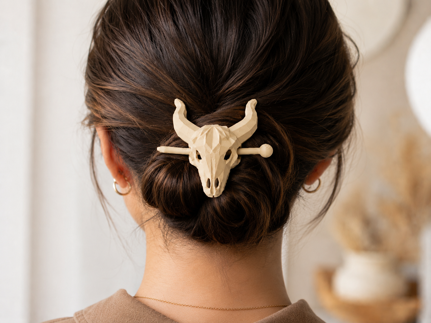 A woman’s dark hair is styled in a bun and held with the Mirabilia Boutique Longhorn Skull Hairpin | Western Theme Hair Accessory in beige. She pairs it with small gold hoop earrings and a brown top, against a softly blurred background.