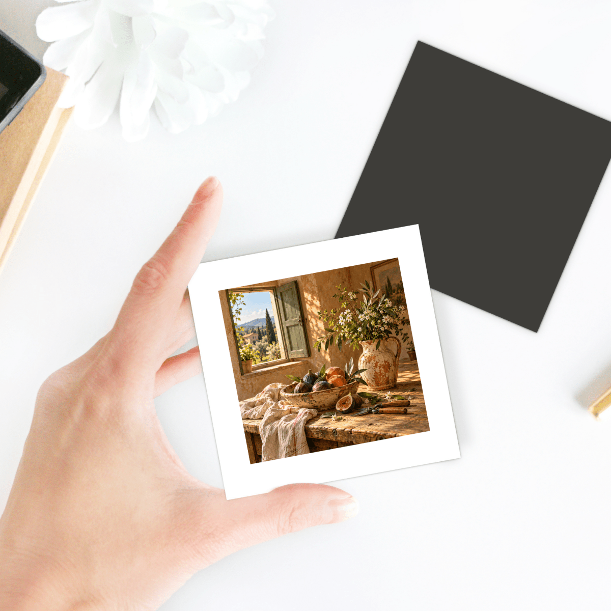 A hand holds a square What the Garden Brought Art Magnet | Ochre & Cypress, featuring a Tuscan still life of figs and a floral vase on a wooden table near an open window.