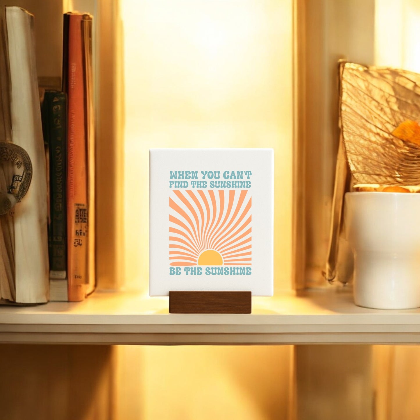 The Be the Sunshine Mini Canvas – Retro Sunburst Sign by Mirabilia Boutique features an orange sun and rays with the quote "When you can't find the sunshine, be the sunshine," glowing warmly amid books and a white pot with golden decor.