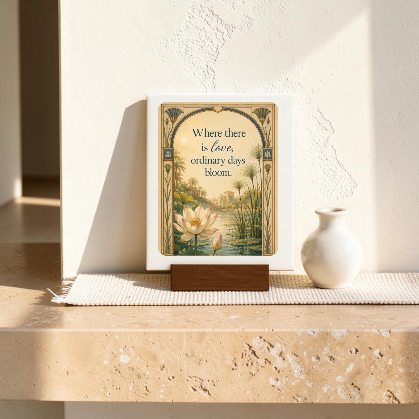 From the Mirabilia Boutique, this “Where There Is Love | Lotus Mini Sign” from the Lapis & Lotus Collection features a delicately framed lotus flower and the words “Where there is love, ordinary days bloom.” Displayed on its wooden stand beside a white vase, it rests on a light stone shelf illuminated by sunlight.