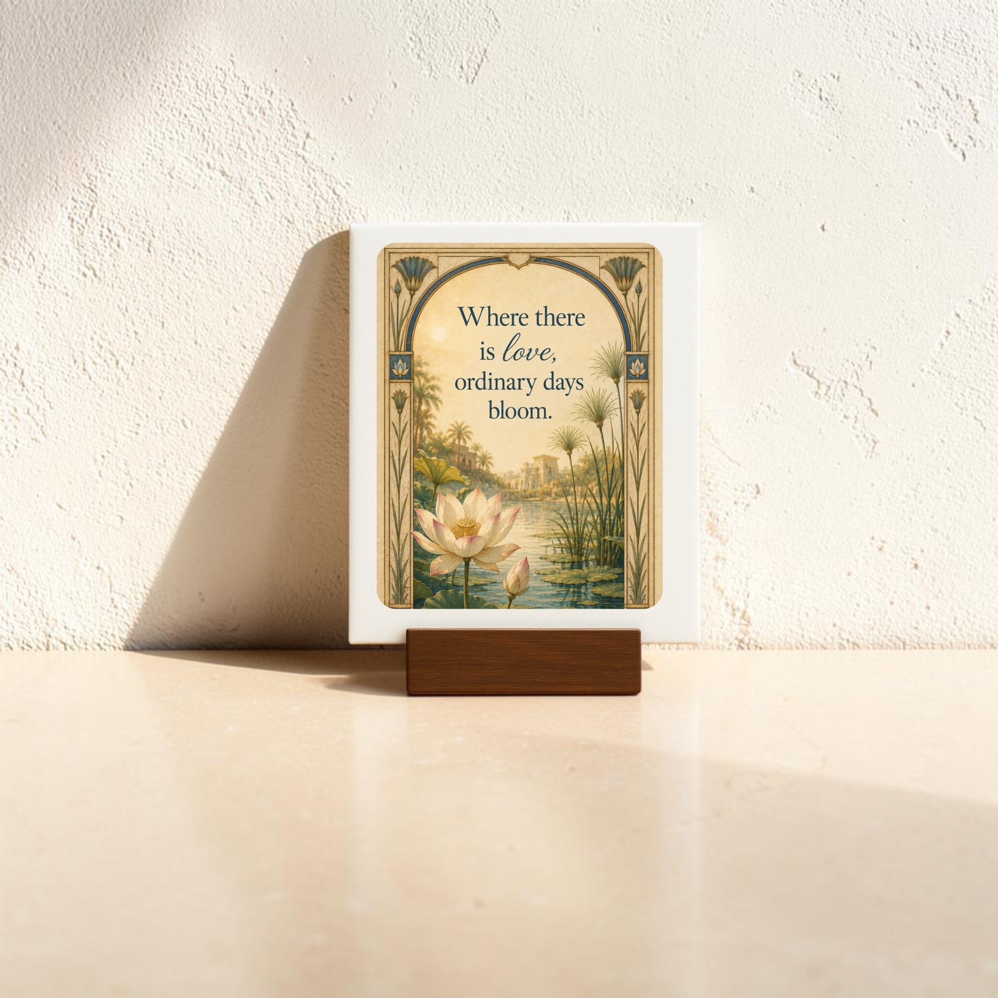 The Where There Is Love | Lotus Mini Sign by Mirabilia Boutique features a lotus flower and reeds alongside the phrase, “Where there is love, ordinary days bloom.” This charming piece from the Lapis & Lotus Collection is displayed on a small wooden stand against a sunlit textured wall.