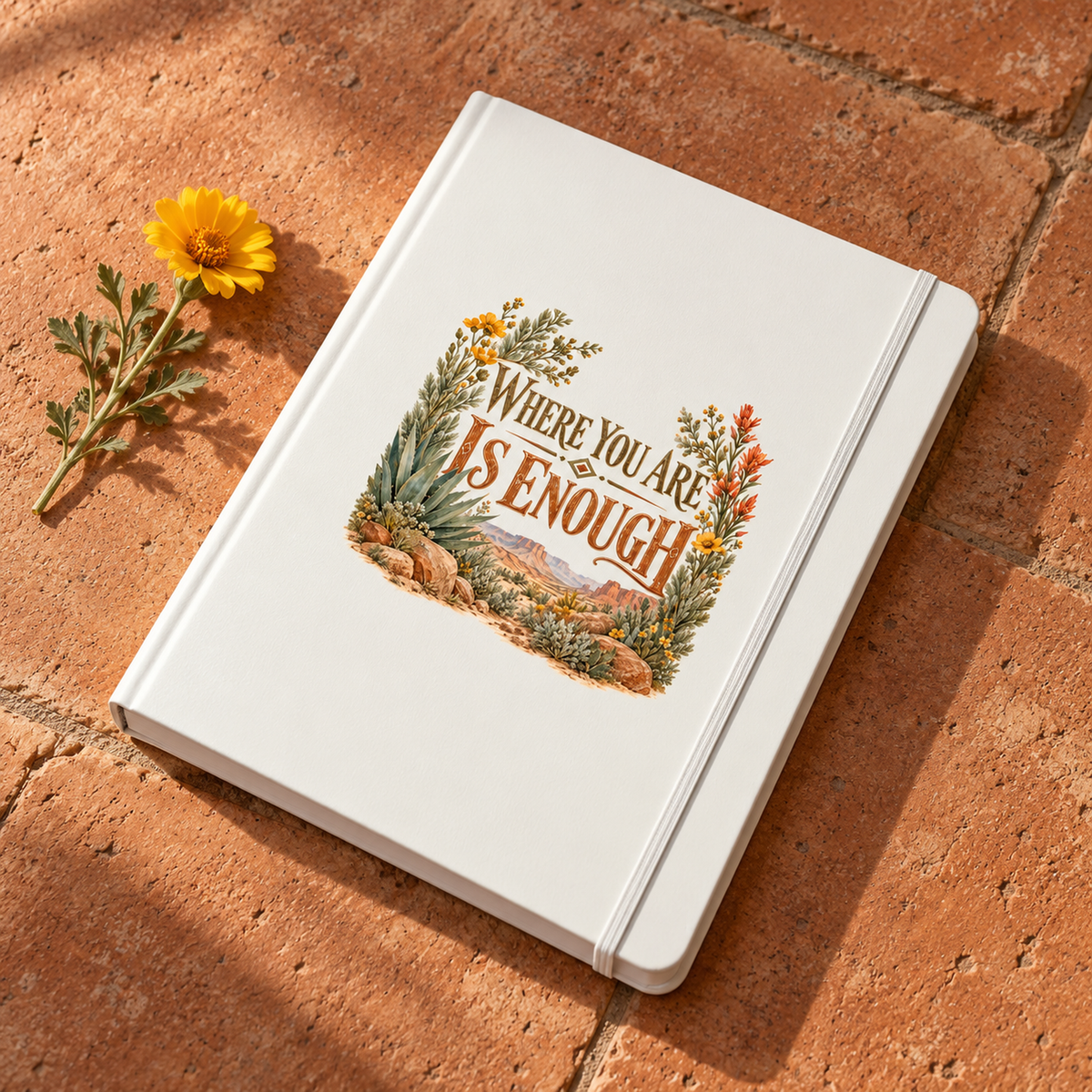 The Where You Are Is Enough A5 Lined Journal | Sage & Stone by Mirabilia Boutique, featuring a white floral cover, sits on terracotta tile beside a yellow flower.